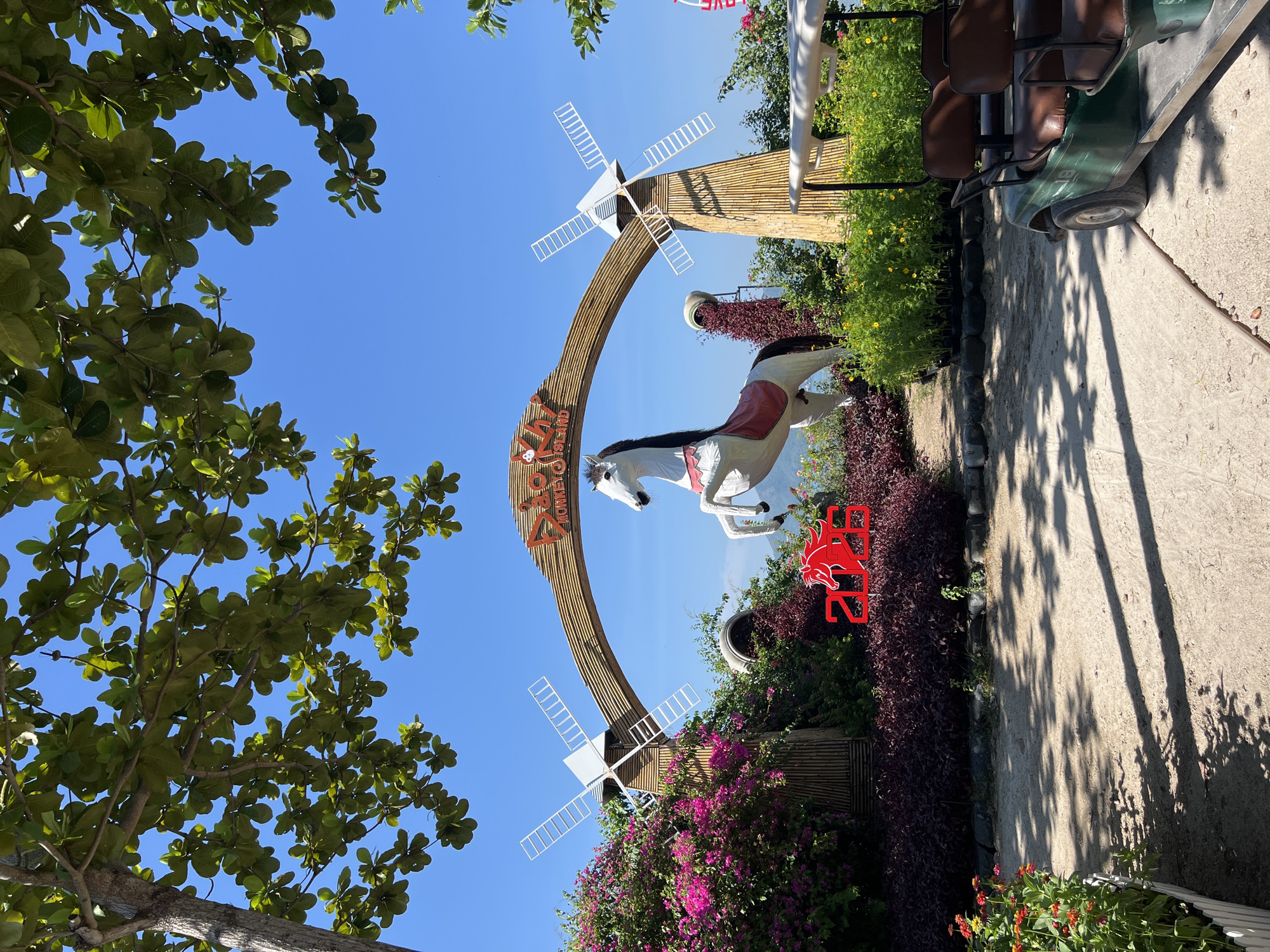 The image depicts an outdoor scene with a clear blue sky and lush green foliage. Prominently featured is a large, decorative archway made of bamboo, with the text "DIO MONKEY ISLAND" visible on it. A white horse statue is positioned under the arch, appearing to rear up. On either side of the arch are windmill-like structures. The foreground is filled with various plants, including vibrant pink and purple flowering bushes, dark red foliage, and smaller yellow flowers. Red numbers "2026" with a horse silhouette are displayed in the lower center of the image. To the right, a green golf cart with brown seats is parked. The ground is sandy and dappled with shadows from overhead trees. The location is identified as Dio Monkey Island in Ninh Ich City, Vietnam.