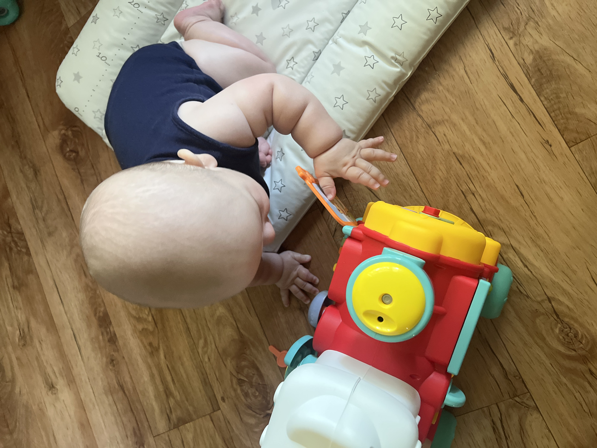 An overhead shot shows a baby, wearing a navy blue onesie, on its hands and knees on a wooden floor. The baby is crawling towards a red, yellow, and white toy train. The train has blue and green accents and a yellow circular button. The baby's hands are reaching out towards the toy. Next to the baby is a light-colored mat with grey stars. The scene is an indoor environment, likely a nursery or play area. The image is taken at an eye-level perspective of the baby.