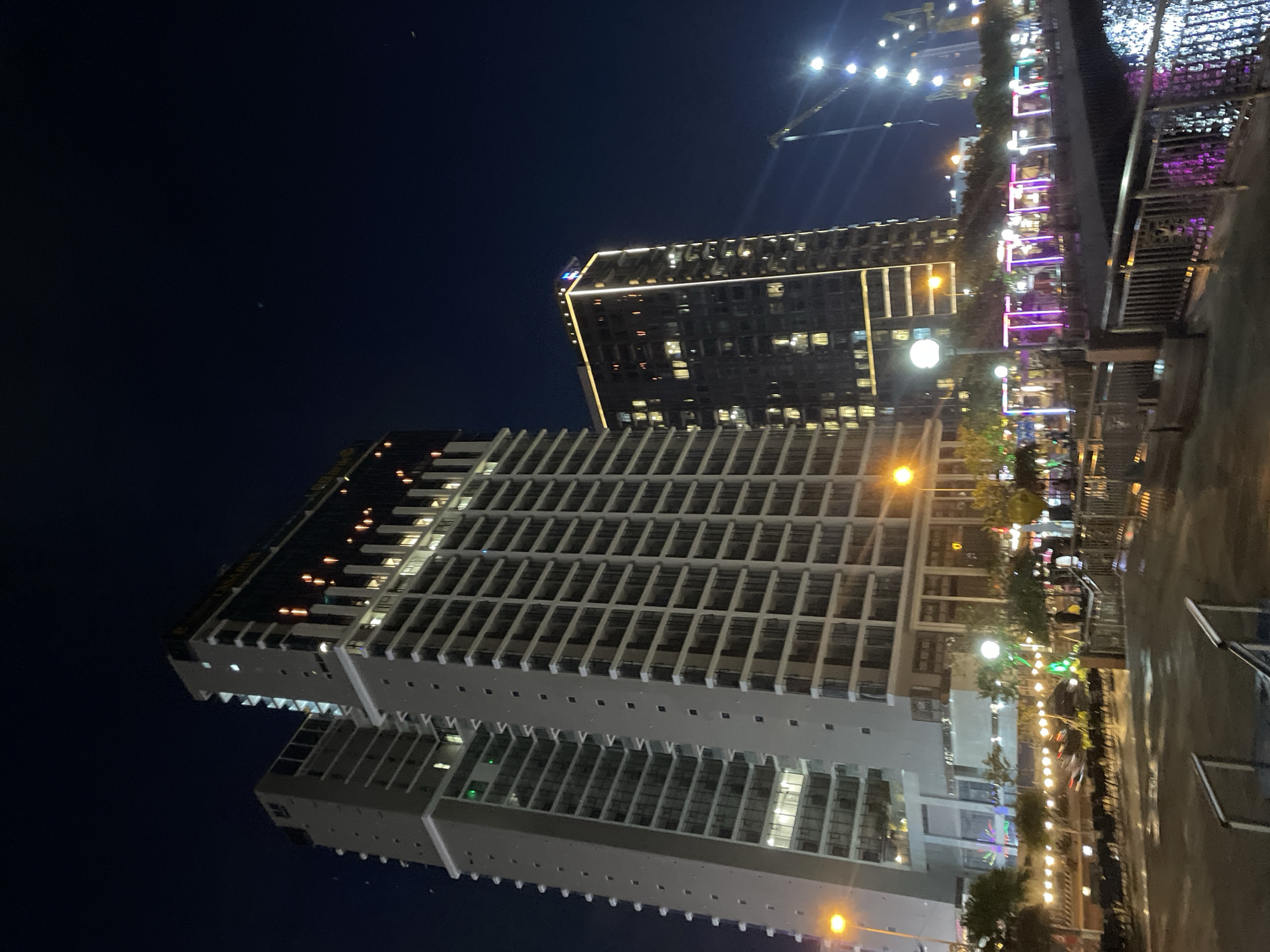 This media file captures a nighttime scene in Đà Nẵng, Vietnam, likely a waterfront area given the presence of illuminated buildings and what appears to be a promenade or walkway. The focus is on two modern high-rise buildings, one significantly taller and more angular than the other, both adorned with exterior lighting that outlines their structures. The taller building features a striking, illuminated crown at its apex.
Below the buildings, a vibrant scene unfolds along a strip of activity. Numerous strings of warm-toned lights illuminate trees and create an inviting atmosphere, suggesting a commercial or entertainment district. These lights are complemented by colorful neon or LED lights, primarily in purple and pink, decorating what appear to be outdoor seating areas or stalls. Some individuals are visible, appearing as silhouettes or blurred figures, engaged in activities typical of a public space at night, such as socializing or dining. The presence of streetlights further enhances the illumination of the area.
The overall impression is one of a bustling urban environment at night, characterized by modern architecture and lively public spaces. The time of day is clearly night, with a dark sky serving as the backdrop. There are no overt indicators of weather conditions, but the reflective surfaces of some areas might suggest recent rain or a damp environment. No specific text is legible within the image. The scene conveys a sense of urban energy and nighttime entertainment.
