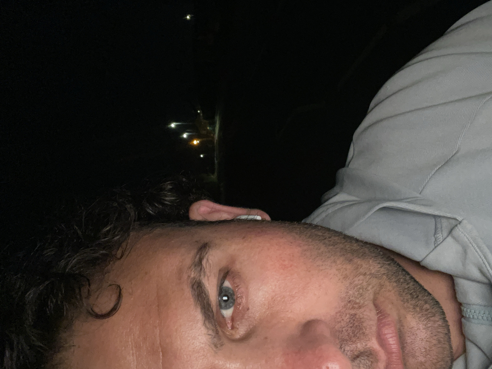 A close-up, low-angle, outdoor, nighttime, selfie-style photograph is captured from the neck up of a person. The subject's left eye, nose, mouth, and part of their ear and hair are visible, positioned on the left side of the frame. The person has light blue eyes, dark, curly hair, and stubble. They are wearing a light blue zippered hooded sweatshirt. An earbud is visible in the subject's left ear. The background is dark and out of focus, with several streetlights visible in the distance, suggesting a road or pathway in an urban or suburban setting within Armagh, United Kingdom.