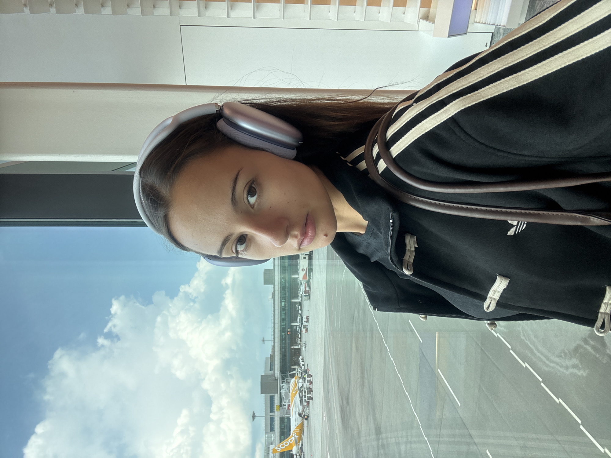A young woman is depicted in a selfie, likely taken at Singapore Changi Airport. She wears headphones and a black Adidas jacket with white stripes. Outside the window, an airport tarmac is visible, with aircraft belonging to the airline Scoot. The sky shows partly cloudy conditions. The buildings of the airport are visible in the background.