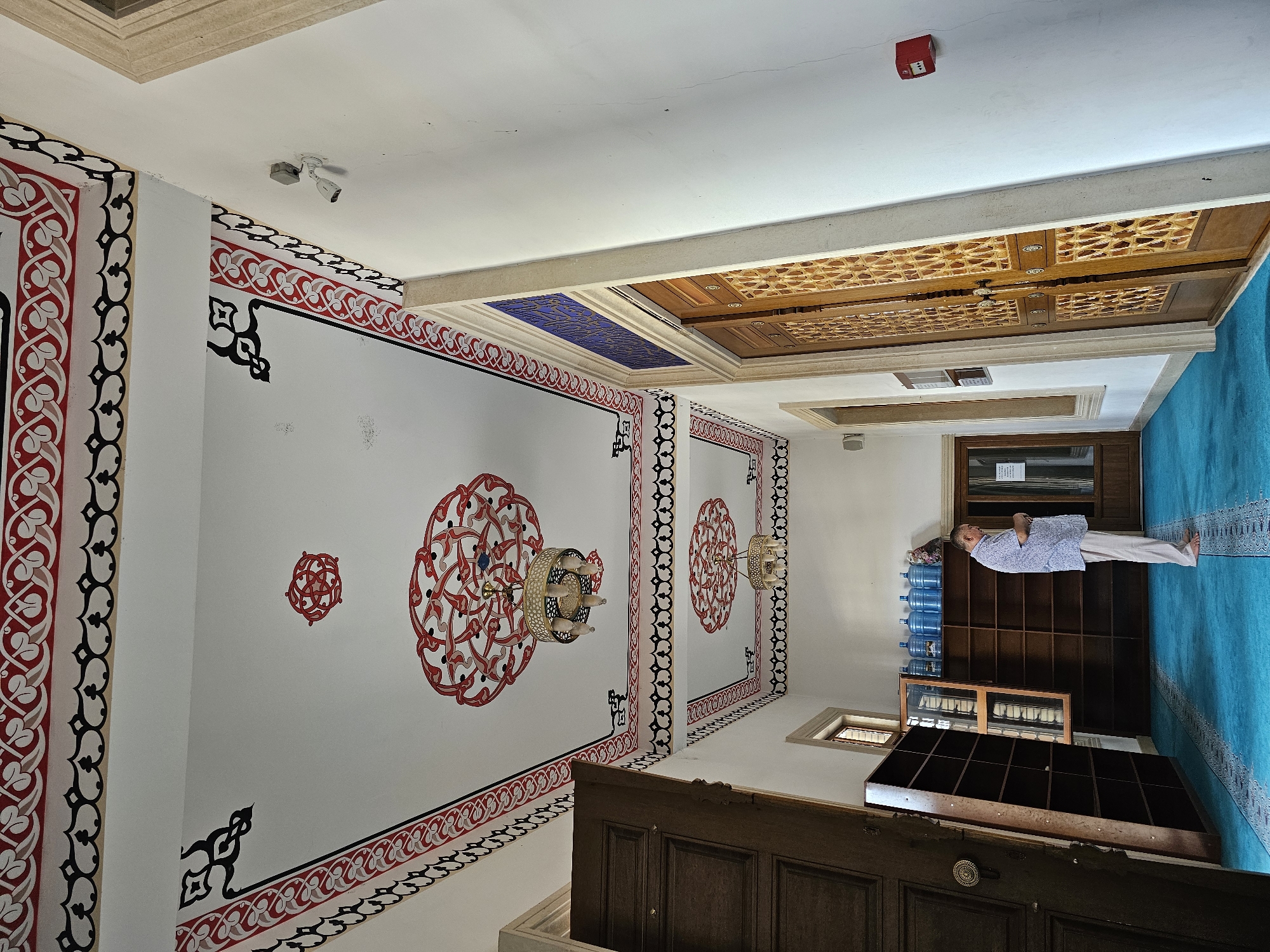A man is standing in a hallway with a blue carpet, facing a wooden door. The hallway has white walls with decorative red and black patterns on the ceiling. There are wooden shelves with bottles on them to the left of the man. To the right of the man, there is another wooden door. The ceiling has two circular chandeliers. The man is wearing a light-colored shirt and pants.