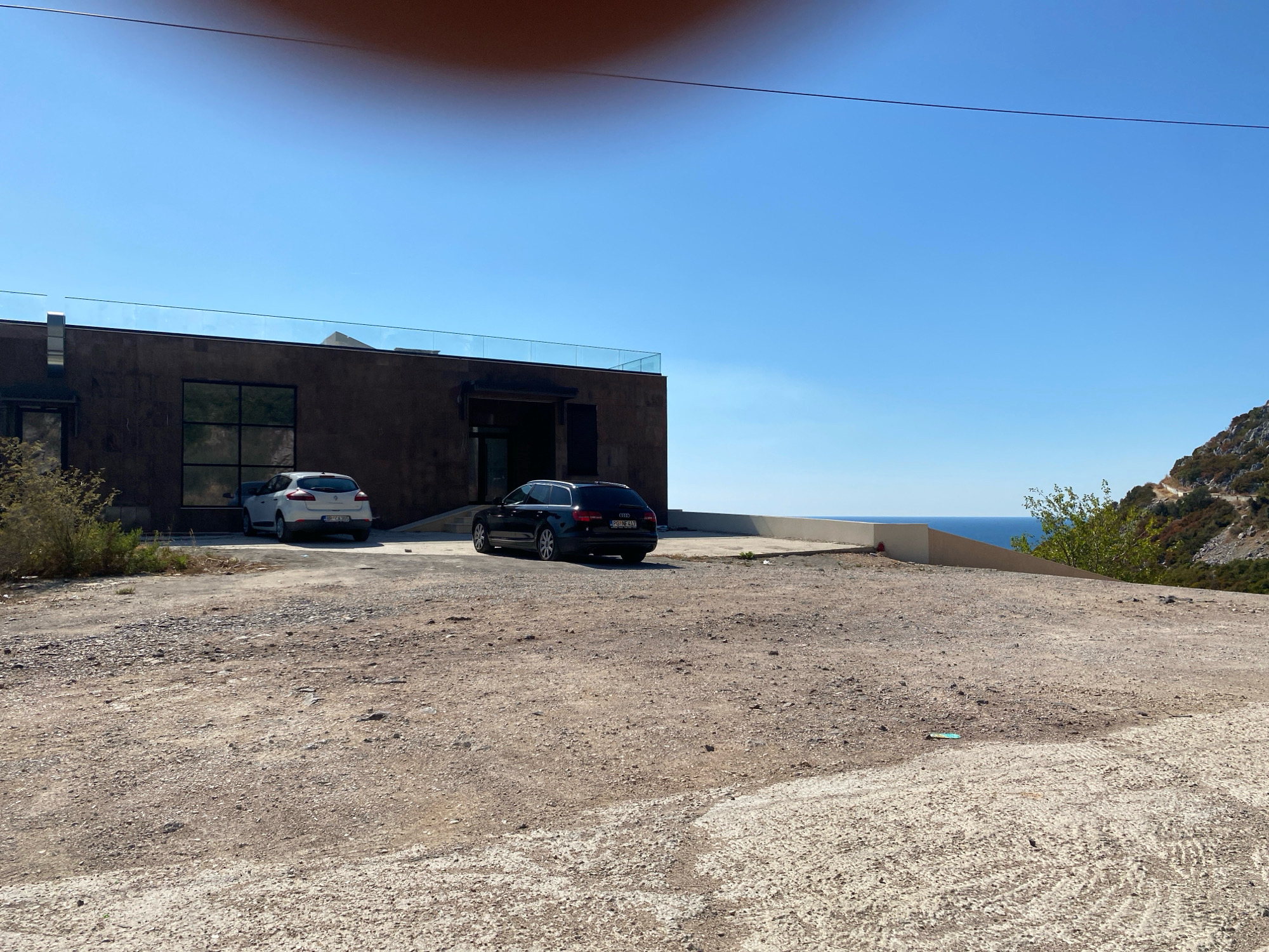 The image shows a modern, dark-colored building with a glass railing on its rooftop, situated in Dobra Voda, Montenegro. In front of the building, on a gravelly, uneven surface, are two parked cars: a white hatchback and a black station wagon. The background features a clear blue sky and a body of water, likely the Adriatic Sea, visible in the distance. The terrain slopes upwards towards a rocky, vegetated hillside to the right of the building. The scene appears to be an exterior view of a property, possibly a residential or commercial establishment, in a coastal environment.