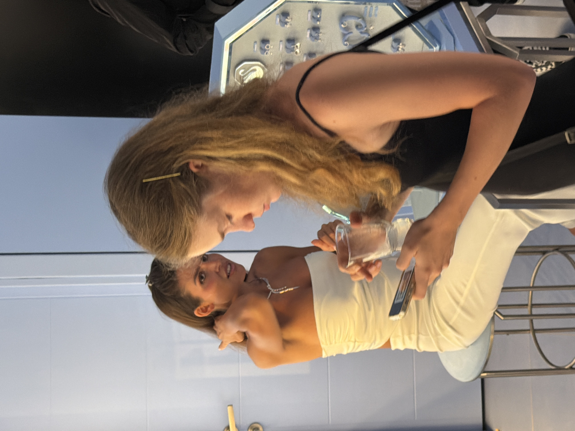The image shows two young women interacting in what appears to be a retail setting, possibly a jewelry store, given the display case of items in the background. One woman, with long blonde hair, leans over and appears to be showing or explaining something to the other woman. She is holding a small, clear object, possibly a sample or a piece of jewelry, in her right hand. The second woman, with dark hair pulled back, is looking at the object with interest, her hand resting near her face. She is wearing a strapless white top and a necklace. The background features a brightly lit display case with various jewelry pieces. The overall lighting suggests an indoor environment, likely during daylight hours, though the precise time of day is not discernible. The mood seems casual and engaged, with both women appearing relaxed. The backdrop of pale blue tiled walls and metallic fixtures hints at a modern commercial space. There is no visible text in the image.