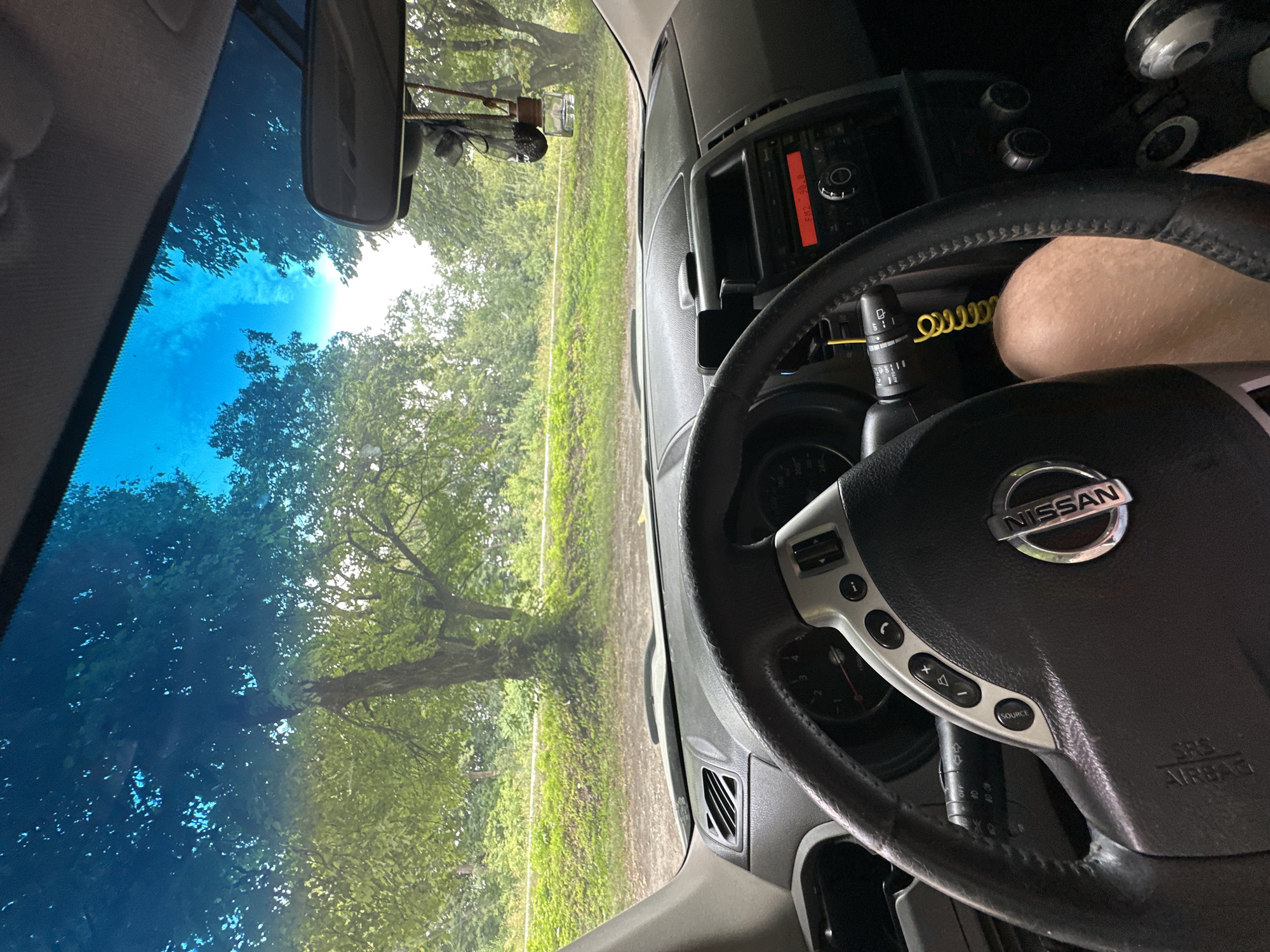 The image is a first-person perspective from inside a Nissan vehicle, looking out through the windshield. The dashboard and steering wheel are in the foreground, with the car's interior features visible, including the radio display showing "FM2 98.8", a yellow coiled cord, and the Nissan logo on the steering wheel. A person's leg is visible on the right side of the steering wheel. Outside the car, a tree-lined road is visible, with lush green trees and foliage under a bright blue sky. The environment appears to be rural or semi-rural. The location of Shatava, Ukraine is not directly evident from the visual information in the image itself; this information is external context.