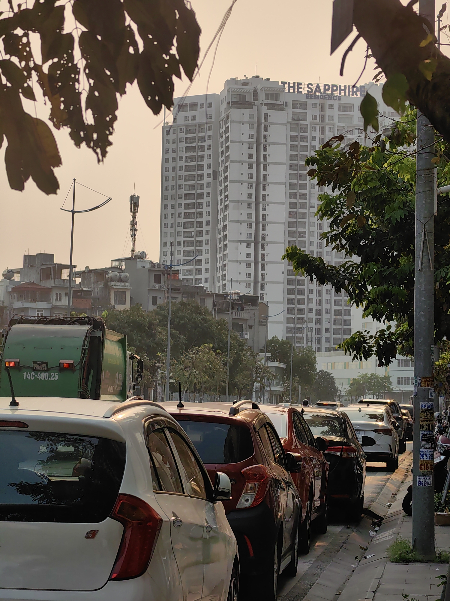 The image depicts a street scene in Hong Gai Ward, Vietnam, characterized by a traffic jam in front of the "The Sapphire Residence" building. Cars are parked along the curb, including a white Kia Picanto, a red car, and a black sedan. A green garbage truck is also visible in the background. Utility poles are present along the sidewalk, with advertisements posted on them. The sky is overcast, with a hazy quality to the light. The architecture consists of a mixture of low-rise buildings and the high-rise residential tower of "The Sapphire Residence." Leaves from trees are visible in the foreground, both in focus and blurred.