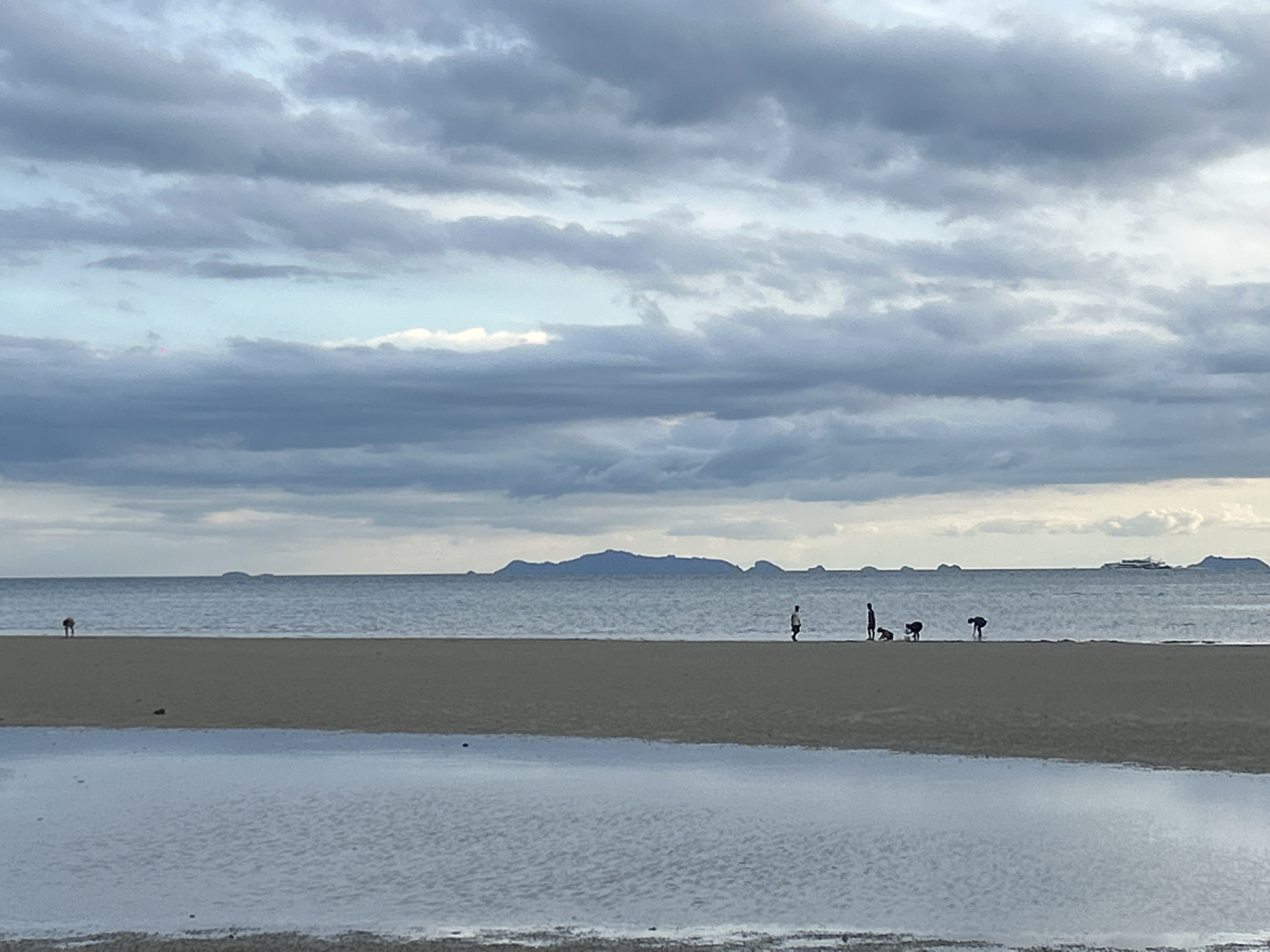 The image depicts a coastal scene at ตำบลเกาะพะงัน, Thailand. Under an overcast sky with grey clouds, a flat, sandy beach is visible in the foreground. A shallow pool of water reflects the sky. Several silhouetted figures are dispersed along the beach. In the distance, the sea stretches out towards an island on the horizon.