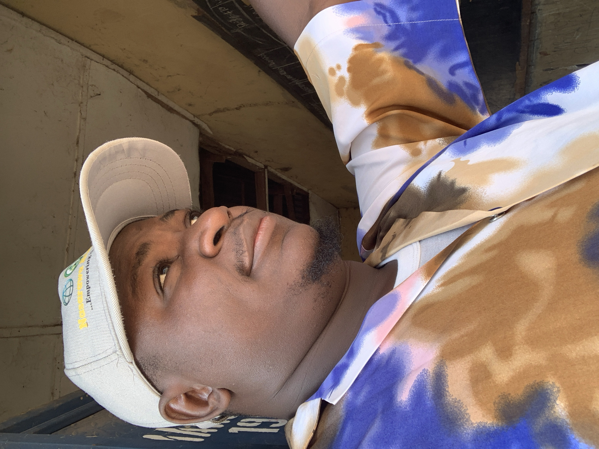 A medium close-up shot shows a man wearing a tan baseball cap and a white undershirt beneath a vibrantly patterned button-up shirt in blue, brown, and pink. The man is looking off to his right, with his face turned slightly away from the camera. He has a small goatee and a light mustache. The background suggests an outdoor or semi-outdoor environment, possibly a rural or semi-urban setting in Akwanga, Nigeria, with elements like rough wooden beams and a wall that appears to be a chalkboard or blackboard. The lighting indicates it is daytime.