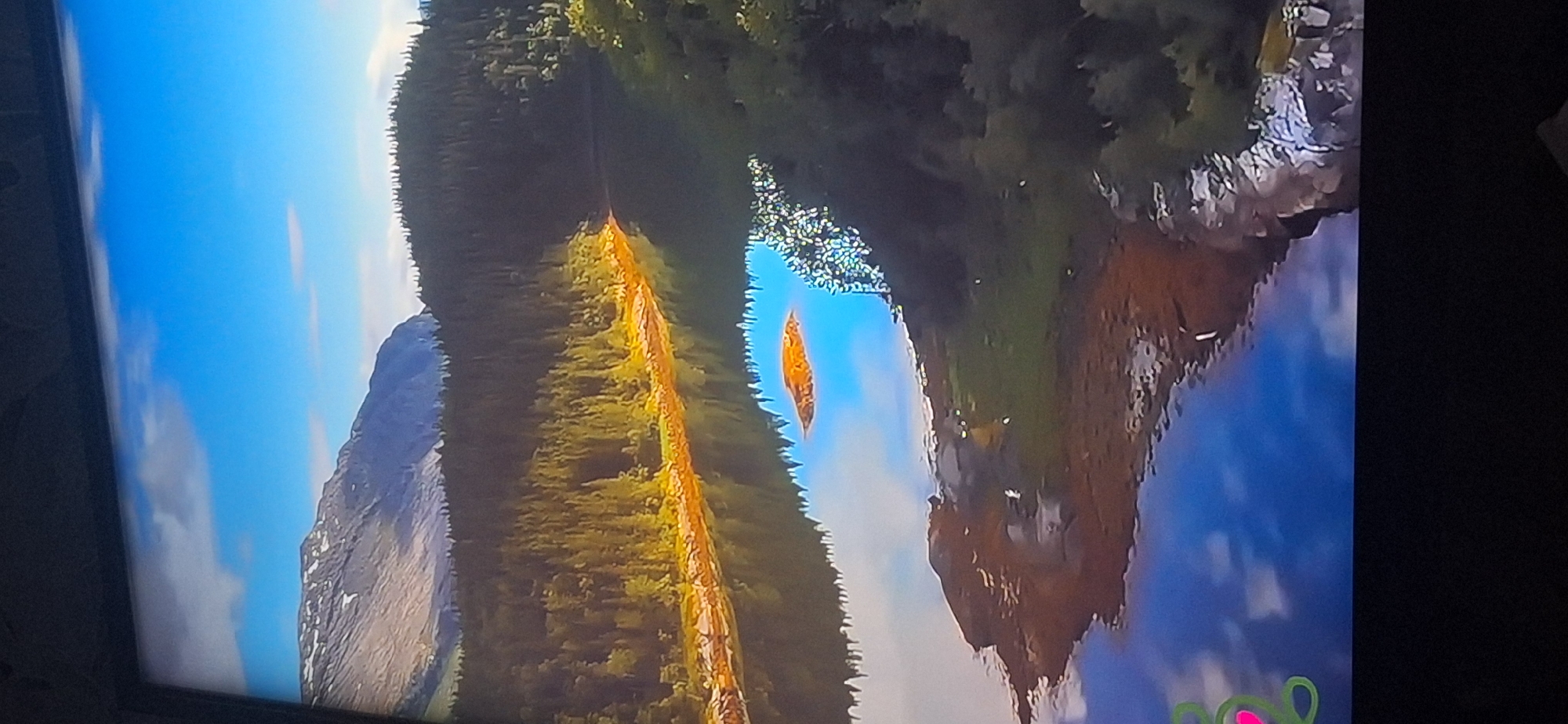 The image shows a scenic landscape with a lake reflecting a blue sky and clouds. Mountains are visible in the background, with a dense forest in the middle ground. The water's edge is lined with trees and rocky terrain. The reflection in the water creates a mirror image of the sky and the surrounding environment. There are no people or structures visible in this image. The scene appears to be a natural, undeveloped area. It is not possible to determine the location within the city of Bila Tserkva, Ukraine based on the provided image.