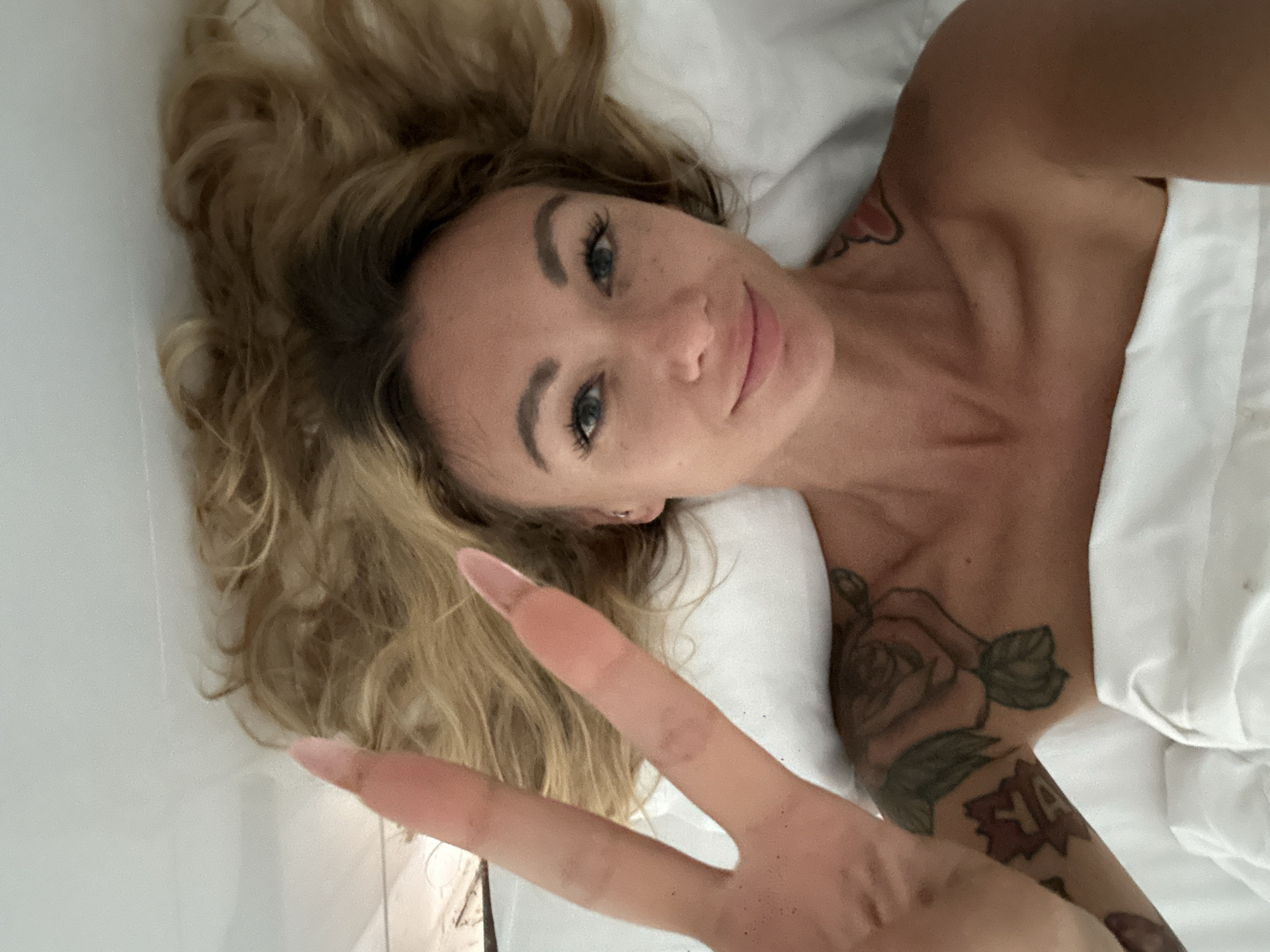 The image is a close-up, overhead shot of a blonde woman lying in bed, looking directly at the camera. Her hair is spread out around her head. She is wearing no visible clothing above the white sheet covering her. Her right hand is raised in front of her face, showing two fingers extended in a peace sign. She has a tattoo of a rose on her left shoulder and upper arm. The background appears to be a white duvet or sheet. The scene is indoors, in what appears to be a bedroom. There is no indication in the image itself to suggest a location in Da Lat, Vietnam.