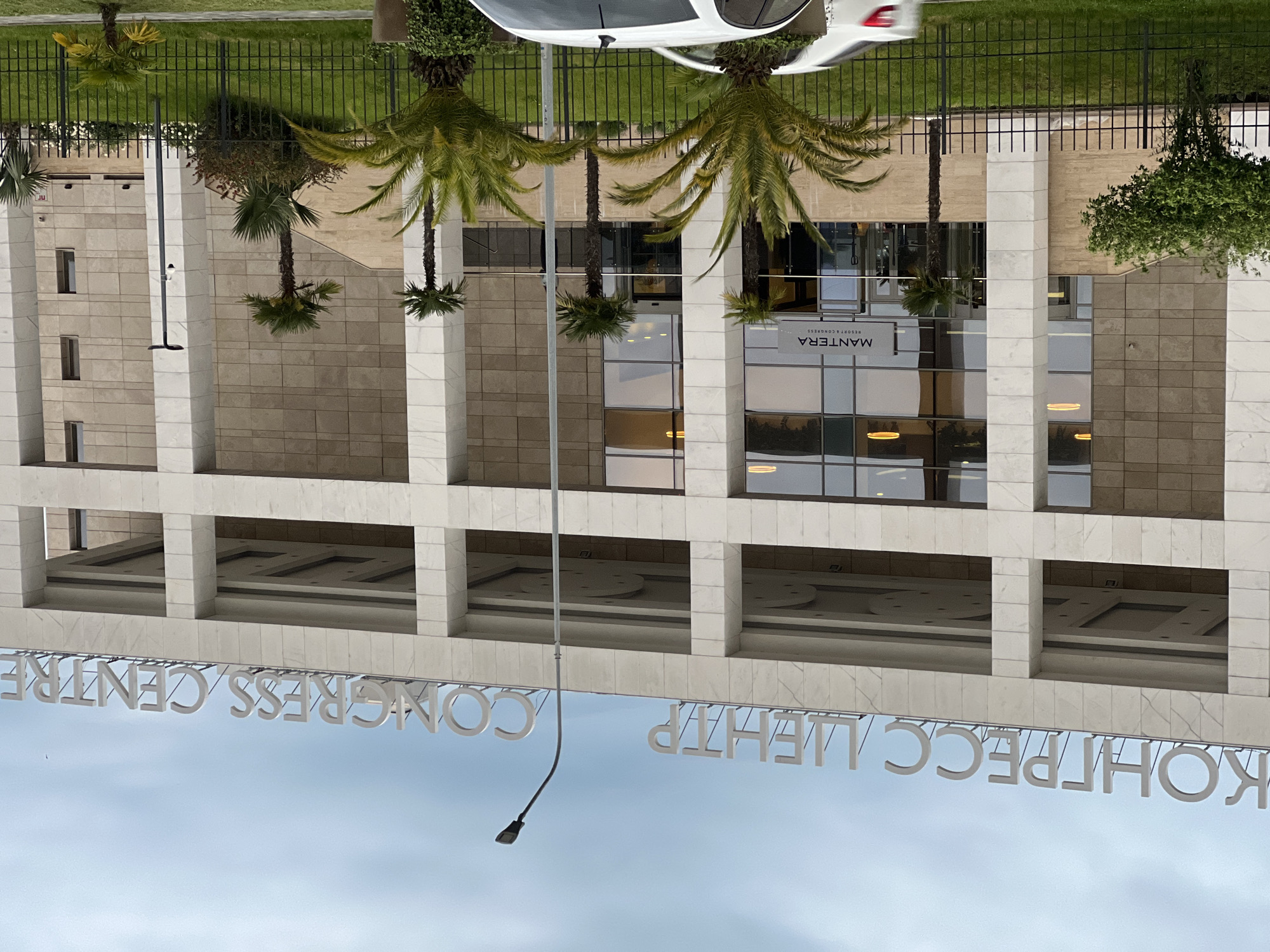 The image depicts the Mantera Resort & Congress Centre in Sochi, Russia. The multi-story building has a modern design with rectangular pillars and a light-colored facade. Large windows reflect the surrounding environment. Palm trees line the entrance area, and a white car is parked nearby. A black metal fence borders the property. A street lamp is visible near the building's sign, which reads "КОНГРЕСС ЦЕНТР / CONGRESS CENTRE." The sky is overcast.
