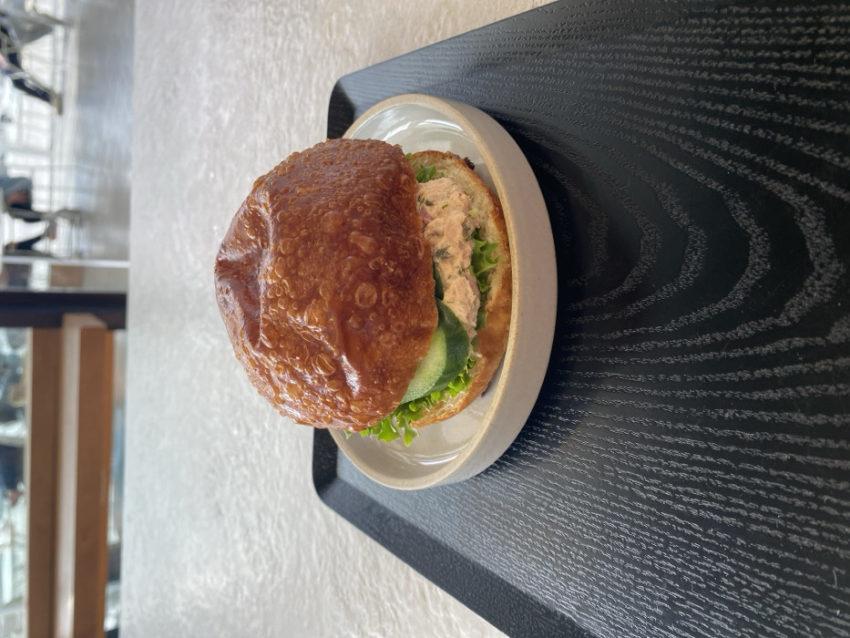The image displays a close-up, slightly elevated view of a sandwich situated on a table within a modern cafe or restaurant in Tel-Aviv, Israel. The central subject is a round, golden-brown bun sandwich with a visibly textured surface, resting on a small, off-white ceramic plate with a narrow rim. The sandwich filling includes light green lettuce leaves, two round slices of green cucumber, and a pale pink, finely textured spread or salad, containing small dark flecks. This plate is positioned on a rectangular black serving tray, which features a prominent diagonal wood grain pattern. The tray rests on a light-colored, subtly textured tabletop. The background is out of focus, showing blurred figures of several individuals, some seated and some standing, along with indistinct architectural elements and furniture, viewed through large windows that suggest an expansive interior or an outdoor view.