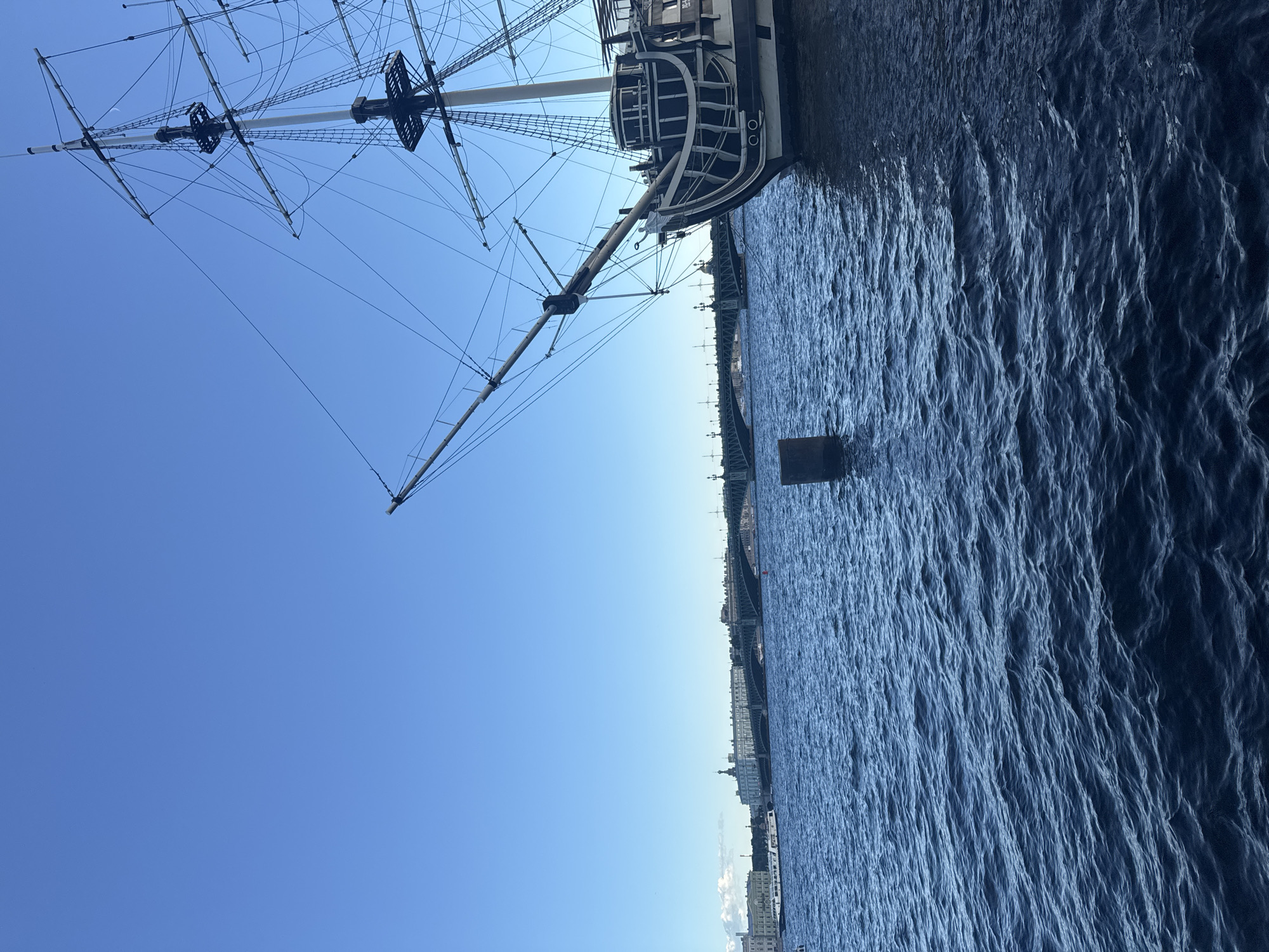 The image depicts a clear blue sky and a deep blue body of water, likely a river or canal. In the foreground, on the right side, is the stern of a historical sailing ship, its dark hull contrasted with white trim. Rigging and masts extend upwards and to the left. In the middle ground, a long, green bridge spans the water, with several buildings visible on the far bank, suggesting an urban setting. A white passenger ship is seen on the water near the bridge. A dark, cylindrical object, possibly a buoy or a mooring post, is partially submerged in the water to the left of the sailing ship. The overall scene suggests a sunny day in a city with significant historical maritime presence. This scene is likely Saint Petersburg, Russia, given the architecture and prominent bridges, possibly near the Neva River.