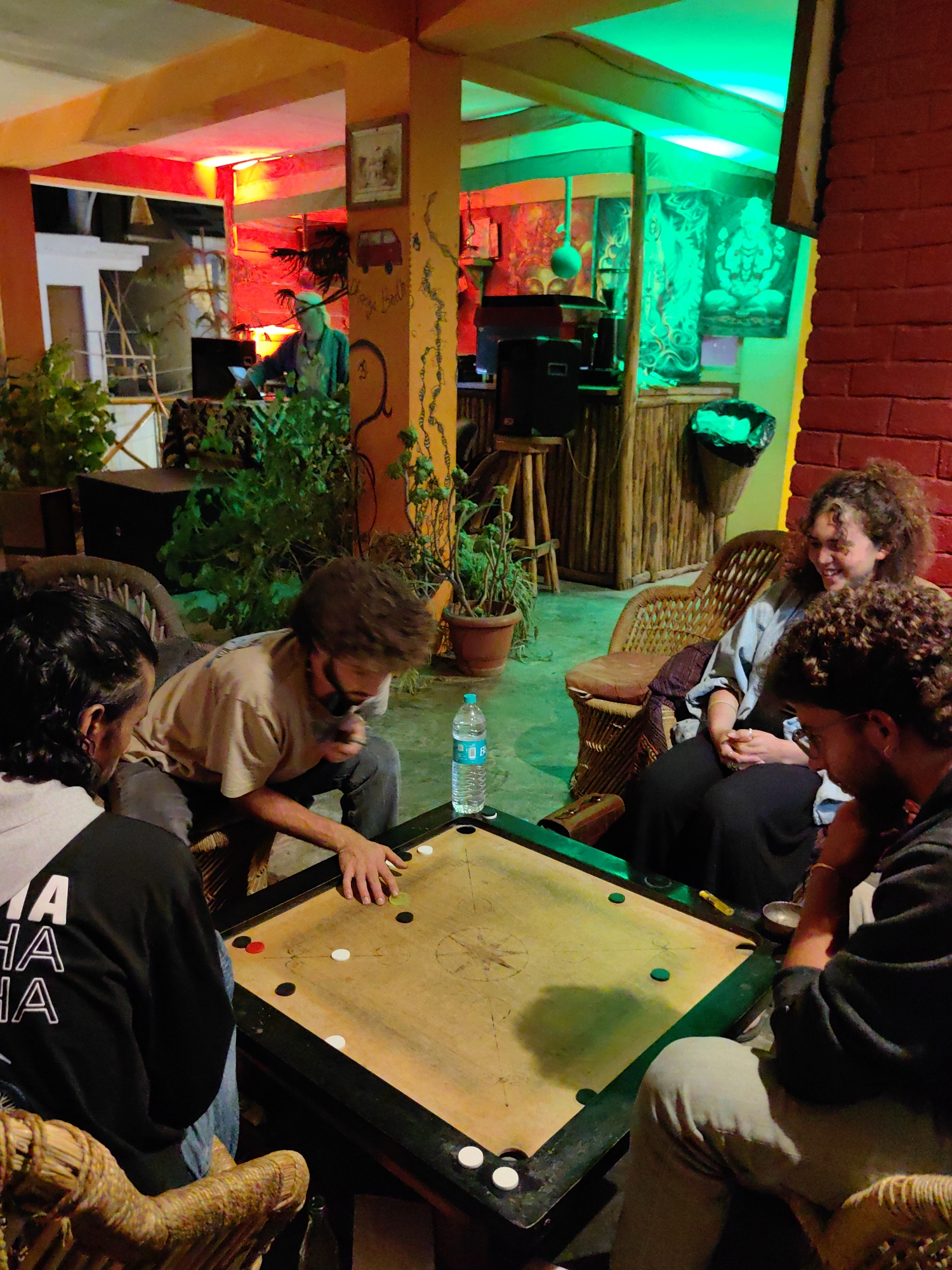 In Vashisht, India, a group of four individuals are gathered around a carrom board in a brightly lit, indoor/outdoor space with vibrant red and green lighting. The scene appears to be a casual hangout, possibly a cafe or common area. In the background, a person wearing a light green cap and shirt is visible behind a DJ setup, with speakers and a laptop. Colorful artwork adorns the walls. The focus is on the carrom game, with two men actively playing and two others observing or waiting their turn. One man is positioned over the board, about to strike a piece with a striker. A bottle of water sits on the table. The ambiance is relaxed, with the warm lighting and the activity of the game creating a social atmosphere.