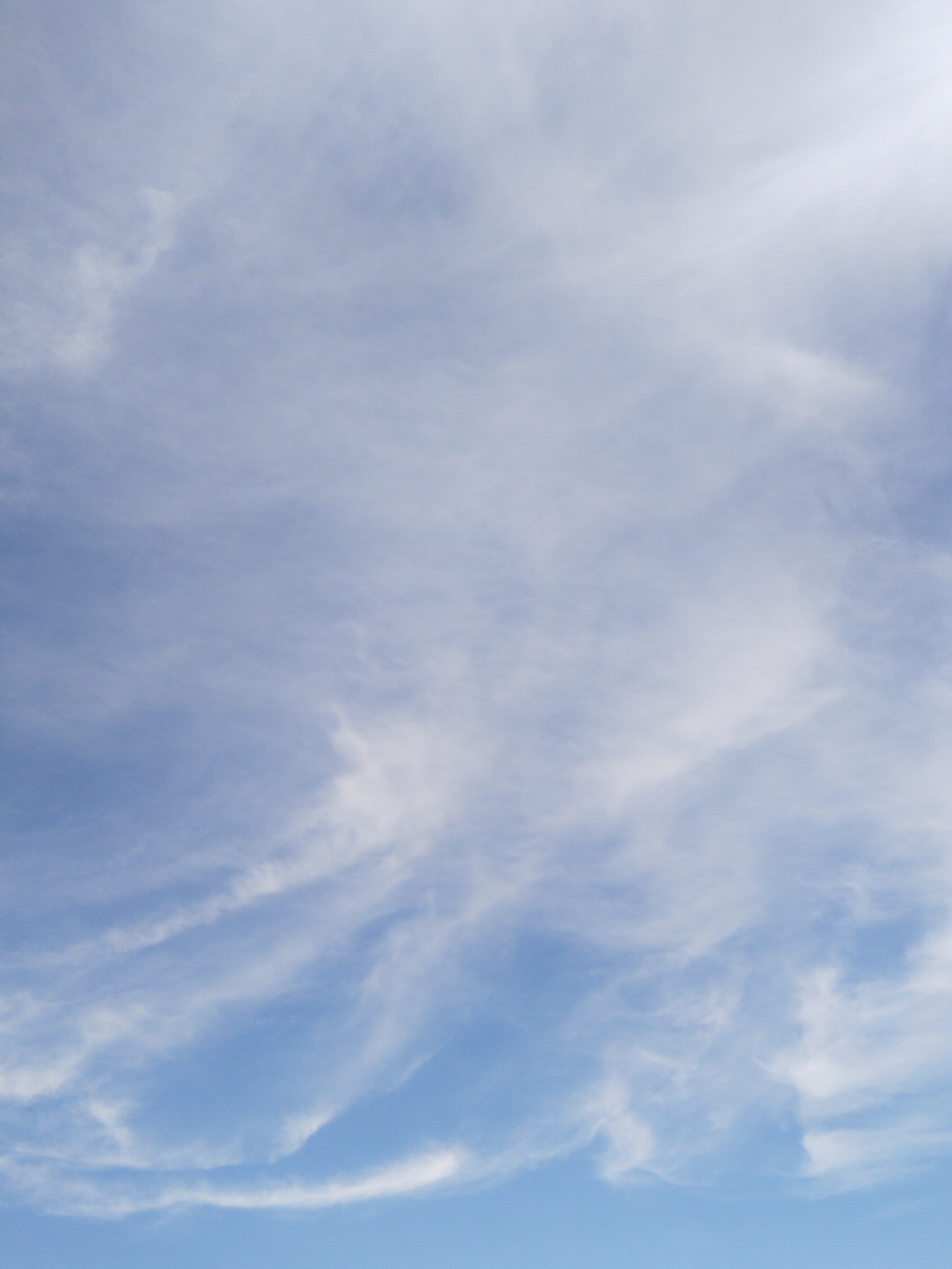 The image shows a clear blue sky with scattered wispy clouds. The clouds are thin and translucent, with some appearing as delicate streaks across the sky. The lower portion of the image is a deeper blue, suggesting a horizon line, though no land features or structures are visible. The lighting indicates it is daytime. There are no people, subjects, or objects in the scene. The environment depicted is solely the sky. The location in the city of Нұресіл, Kazakhstan is not ascertainable from the image content.