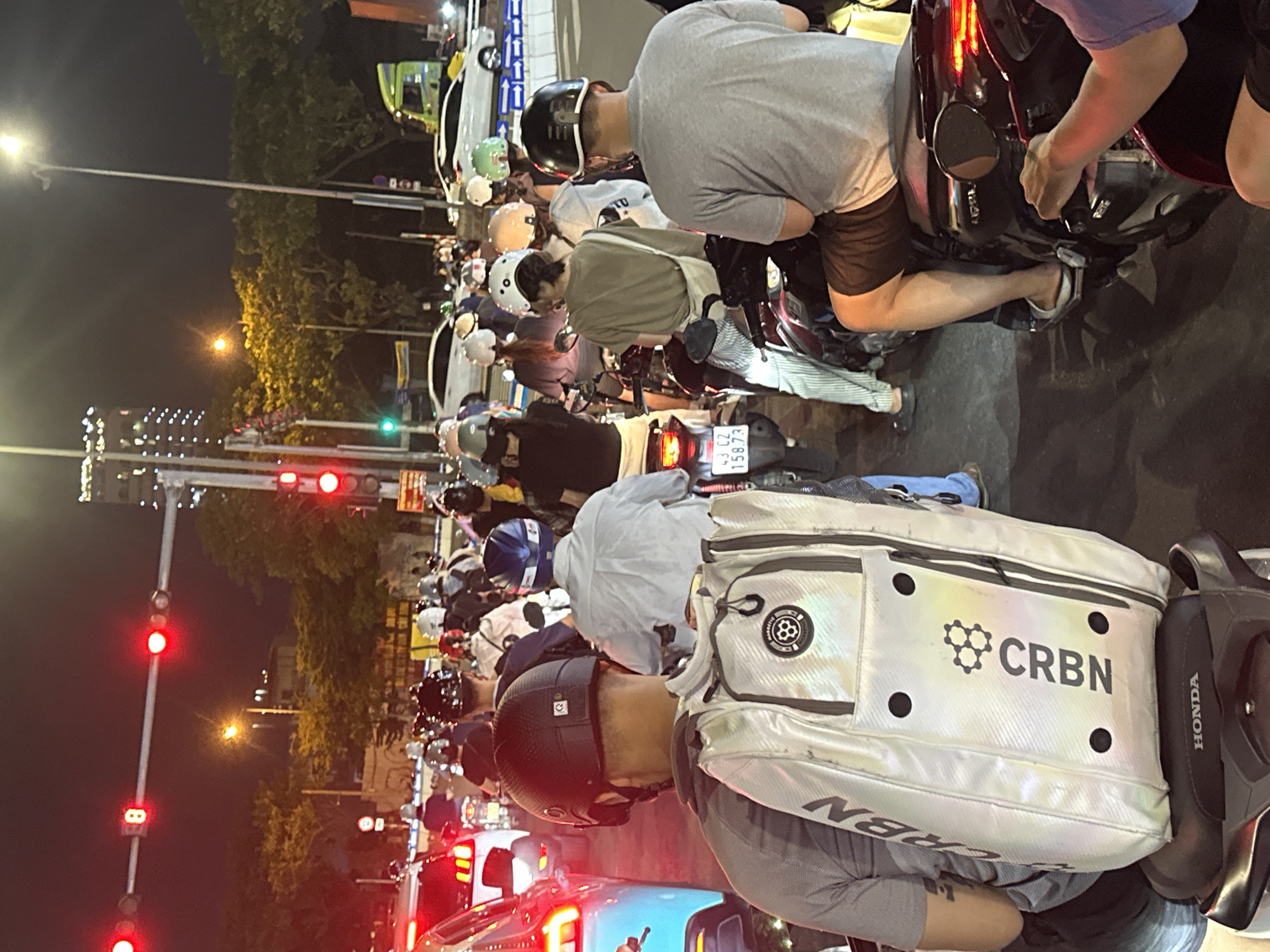 The image depicts a nighttime street scene in Da Nang, Vietnam, likely at an intersection with traffic lights. Numerous people on motorcycles are stopped, suggesting a traffic jam or waiting for a signal change. The overall impression is one of urban life and congestion.
The main subjects are the people riding and waiting on their motorcycles. They are wearing various casual clothes and helmets, some of which are brightly colored. Visible objects include the motorcycles themselves, streetlights, traffic signals displaying red lights, and a silver bag with the brand name "CRBN" prominently displayed. There's also a Honda logo visible on a motorcycle part. The setting is an urban street at night, illuminated by artificial lights and the red glow of traffic signals.
The activity taking place is primarily waiting in traffic. The individuals appear calm, suggesting this is a common occurrence. The time of day is clearly night, indicated by the dark sky and artificial lighting. While weather conditions aren't explicitly shown, the lack of rain and the general atmosphere suggest it might be a clear night. The location cues, such as the Vietnamese license plate "43 C2 158.73", help pinpoint the location to Da Nang, Vietnam. The overall mood is ordinary and everyday, devoid of strong emotional expressions.