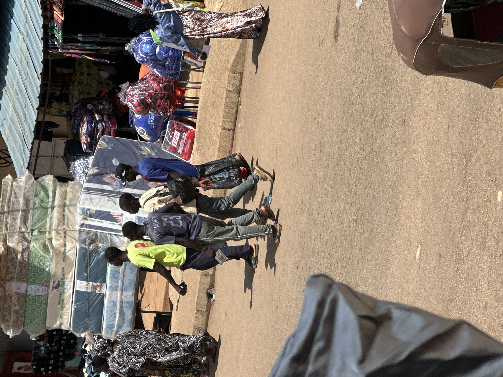 In Bukuru, Nigeria, a street scene unfolds with a group of individuals walking along a dusty asphalt road. In the foreground, a black plastic bag, partially obscuring the view, lies on the ground. Behind it, a brown auto-rickshaw is partially visible on the right side of the frame.
A woman in a patterned dress and headscarf walks from left to right. Following her are four young men. The first, wearing a bright green t-shirt and dark pants, walks with a phone in his hand. Next to him, a man in a dark blue t-shirt with the number "23" on the back and gray striped pants walks, holding a small packet. Another man in a cream-colored shirt and jeans follows. To the right of the group, a woman in a denim jacket and a long, leopard-print skirt walks towards the viewer.
In the background, a market stall is set up. Piled high are mattresses wrapped in plastic, along with various other goods displayed on shelves and racks. The stall has a corrugated metal roof. The environment suggests a busy commercial area in Nigeria.