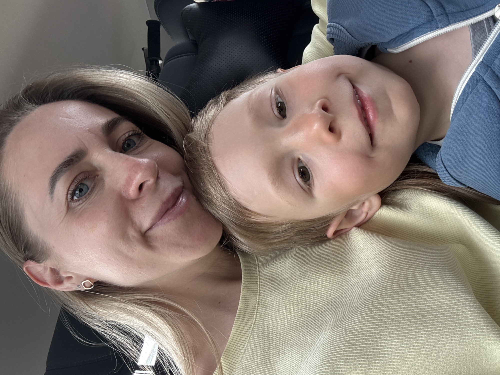 A woman and a child are in close proximity, likely posing for a photograph. The woman has fair skin, blue eyes, and light blonde hair. She is wearing an earring and a yellow sweater. The child has fair skin, brown eyes, and light blonde hair. They are wearing a blue hooded sweatshirt and a yellow sweater. A black leather chair is in the background. The location cannot be determined from the visual information.