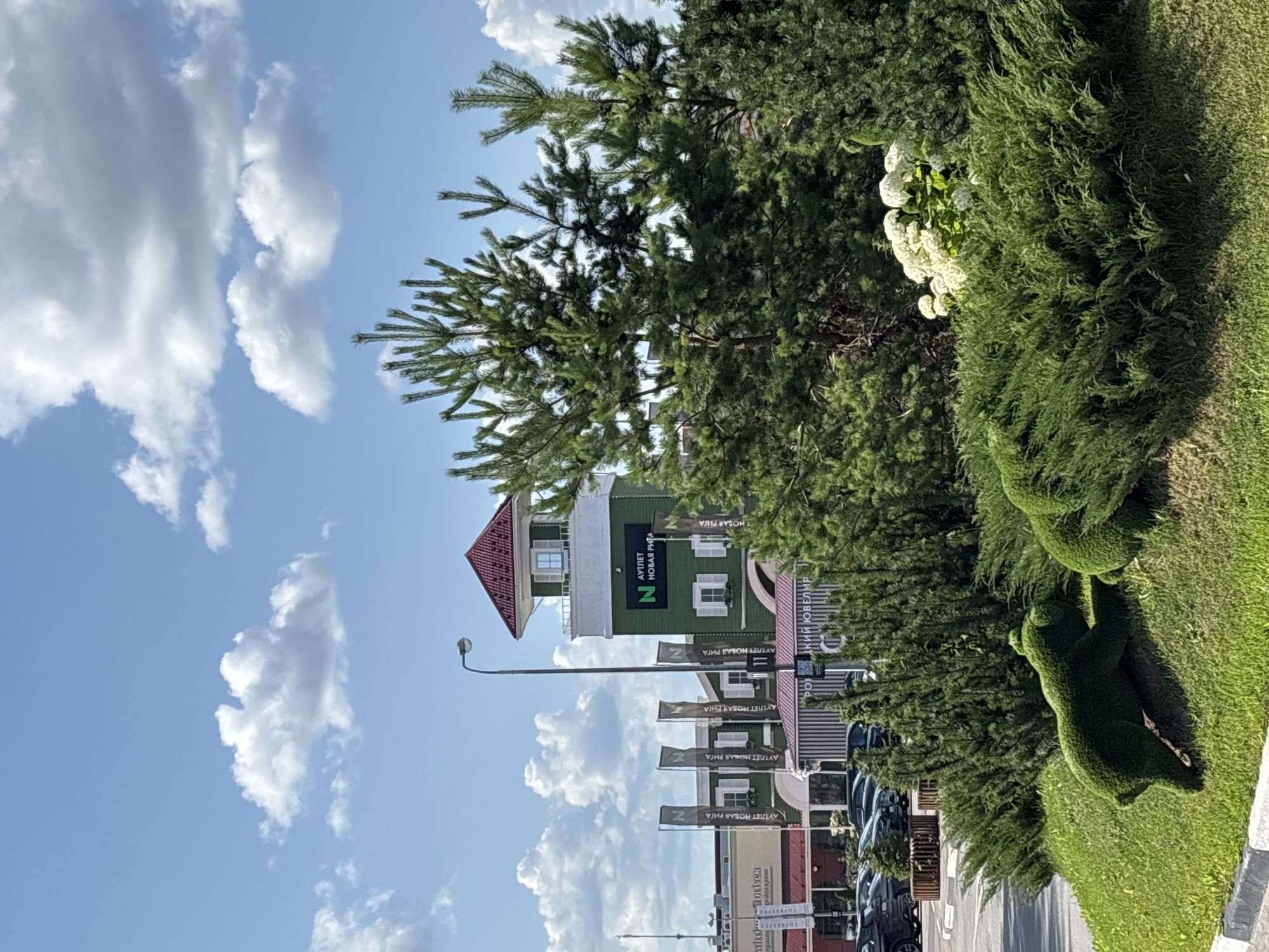 The image shows an outdoor scene on a sunny day with a blue sky and scattered white clouds. In the background, a green building with a red-tiled roof and white trim stands prominently. The building features several windows with white shutters and a tower-like structure at the top. A sign on the building reads "N outlet Nova Riga". Several tall, grey flags with a black logo are positioned in front of the building. To the left of the building, other structures with red roofs are visible, along with parked cars. In the foreground, lush green bushes and manicured grass dominate the landscape. A topiary sculpture resembling a green bear is positioned on a grassy slope. White flowers are clustered amongst the greenery. The scene appears to be part of a commercial area, possibly an outlet mall, in Pokrovskoe, Russia, given the building's signage.