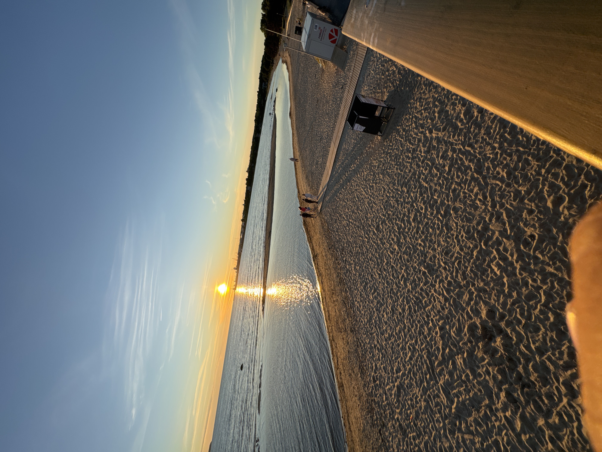 The image depicts a serene beach scene at sunset in Tallinn, Estonia, likely Pirita Beach, known for its wide sandy expanse and views of the Baltic Sea. The sun, a brilliant orb, is low on the horizon, casting a warm, golden glow across the water and sand. The sky transitions from a clear blue at the top to hues of orange and yellow near the horizon, with wispy clouds scattered across it.
The water is calm, reflecting the sunlight in a shimmering path. A gentle curve of sandy beach stretches along the water's edge, marked by footprints and the textured patterns left by receding waves. Several individuals, appearing as small figures in the distance, are walking along the shoreline or gathered in small groups. A wooden boardwalk runs parallel to the beach, providing a path for pedestrians. Adjacent to the boardwalk, a black trash bin and a white kiosk with red signage are visible. The background features a line of trees and, further out, industrial structures hinting at the city's port or industrial area. The overall impression is one of a peaceful, public space enjoying the end of a day.