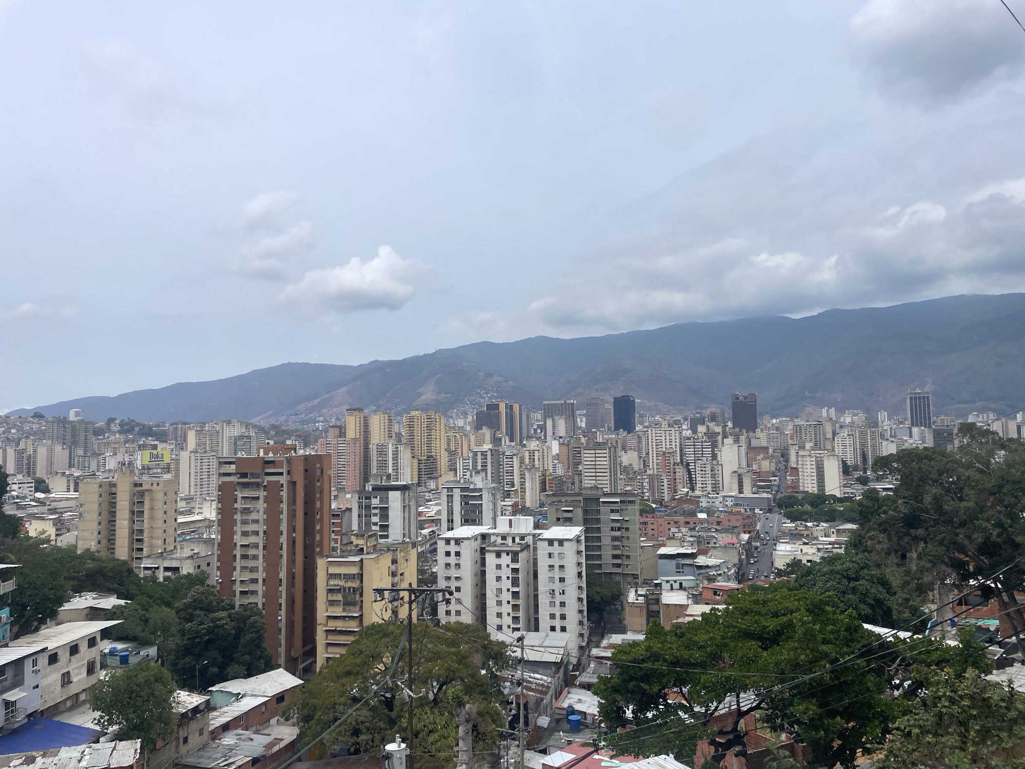 The image shows a high-angle, wide shot of Caracas, Venezuela. The urban landscape is dense, with a mix of high-rise buildings and lower-lying structures extending into the distance. Mountains loom in the background, partially obscured by a cloudy sky. The architecture is varied in style and color. In the foreground, there is vegetation and a view of what appears to be a residential area with rooftops and trees. The sky is overcast. The lighting suggests it's daytime but not brightly sunny.