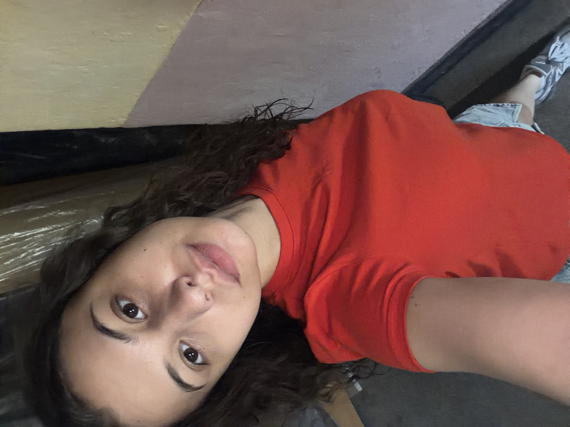 An overhead shot shows a young woman with long, dark, curly hair wearing a bright red t-shirt and denim shorts. Her head is tilted to the left, and she is looking directly at the camera. Her skin is clear and appears smooth, with a few subtle freckles across her nose and cheeks. Her lips are slightly parted, revealing a hint of teeth. The background consists of a wall painted in different shades of yellow and pink, with a dark vertical element on the left. The floor is a dark, textured surface, possibly concrete or asphalt. The woman's right arm is visible, bent at the elbow, and she is wearing a grey and white sneaker on her right foot.
