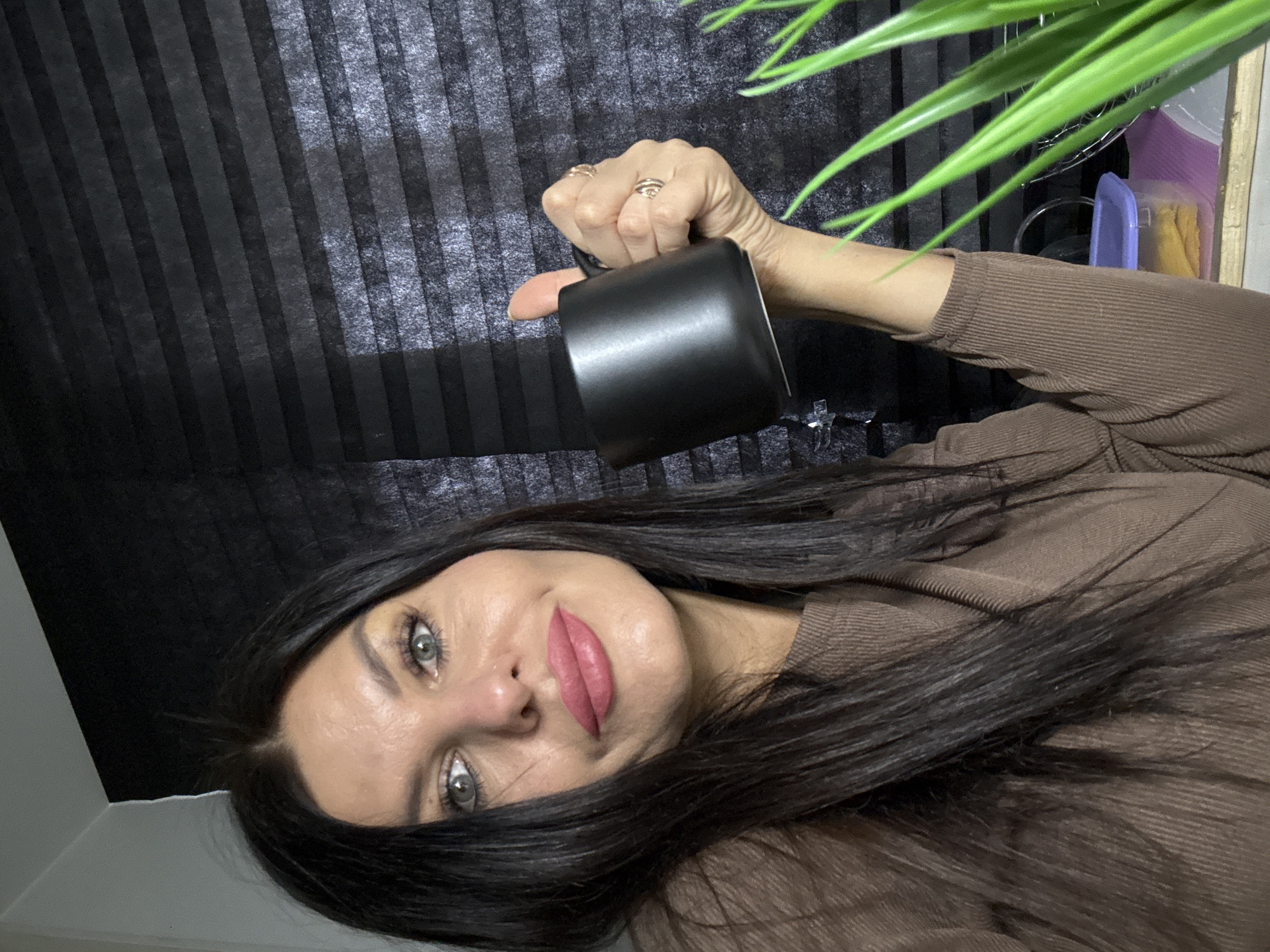 A woman is holding a black mug in front of a black vertically striped wall. She is wearing a brown long-sleeved shirt and rings on her hand. A plant with long green leaves is visible in the upper right corner. The image was taken in a room with white walls. The location of the image is not evident in the city of Chaikovsky, Russia.