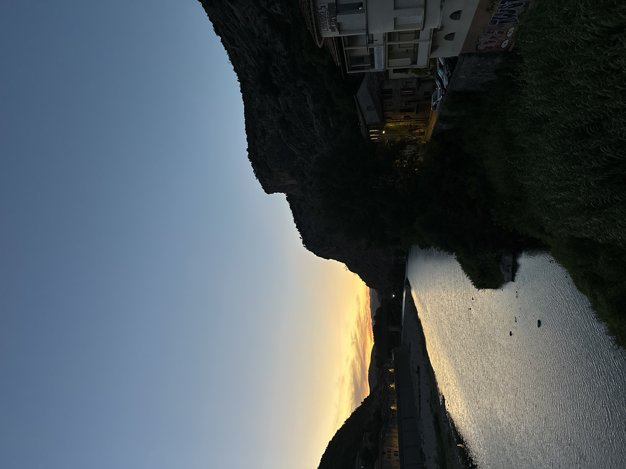 The image depicts a river flowing through a town at dusk. The sky is a gradient of blue and yellow, with the sun having recently set. On the left bank of the river, there are buildings with illuminated windows, suggesting a residential or commercial area. A bridge is visible in the distance, spanning the river. On the right bank, a steep, rocky mountain rises, casting a shadow over the buildings below. One building on the right has a sign that reads "CHAMBRES D'HOTES," indicating it is a guest house. There are several cars parked along the street in front of the buildings. Lush green vegetation lines the riverbanks and slopes. The overall scene is peaceful and scenic, capturing the transition from day to night in a French town.