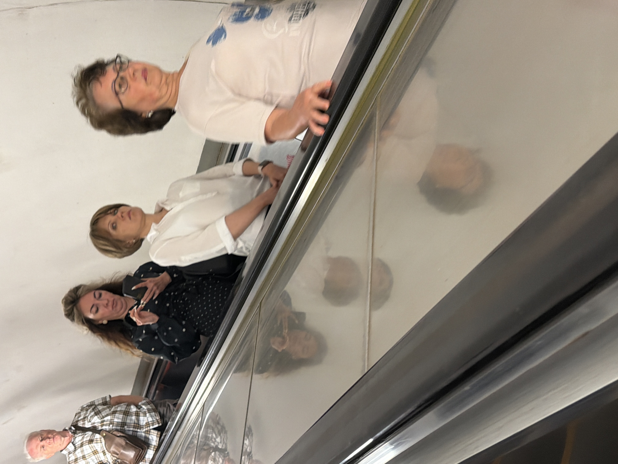 This is a medium shot, eye-level, outdoor photograph of four people on an escalator. The escalator is made of reflective metal and appears to be going upwards. In the foreground, the escalator steps and handrail are visible. On the escalator, from left to right, is a man wearing a plaid shirt and a brown bag over his shoulder, looking straight ahead. Next to him is a woman with long brown hair, wearing a dark polka-dot shirt and holding a smartphone. Further up the escalator is a woman with short brown hair, wearing a white shirt, looking ahead. In the far right of the image is an older woman with short brown hair, wearing glasses and a light-colored shirt, looking to her left. The background is a plain white wall. The overall lighting is bright, suggesting daylight. It is impossible to determine the specific location in Moscow, Russia from the visual information provided.