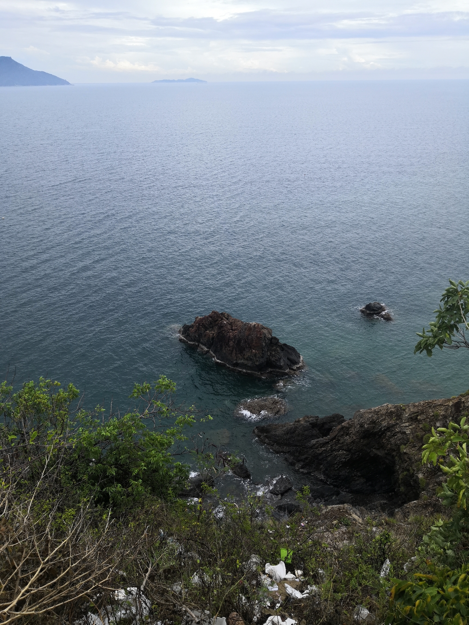 The image depicts a coastal scene in Bac Nha Trang Ward, Vietnam, viewed from an elevated position. The foreground shows vegetation including bushes and some litter. Beyond this, rocky cliffs extend into the water. The sea is visible, with a large rock formation closer to the shore and a smaller one further out. In the distance, landmasses can be seen under a cloudy sky. There are no people visible in the image.