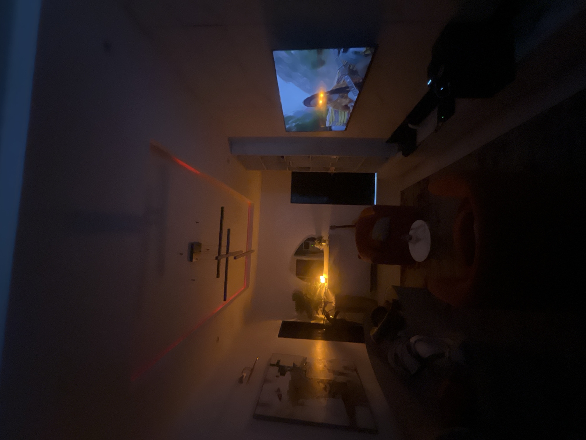 The scene is an interior shot of a room in Igboefon, Nigeria. A television is mounted on the ceiling, displaying a video. Below the TV, there's a shelf unit and a smaller screen. A ceiling fan is visible. A red LED strip is located on the left side of the frame. An orange armchair is present in the room. A floor lamp is turned on and illuminates the lower part of the scene, along with art on the floor. Slippers are near the bottom of the image.