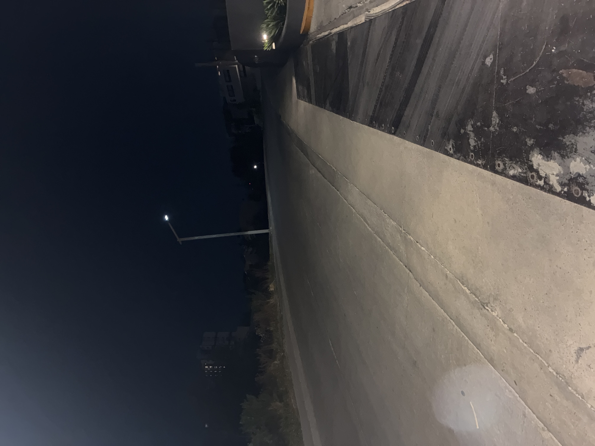 The image shows a street at night in what appears to be the city of Huixquilucan de Degollado, Mexico, based on architectural styles and infrastructure. The street is mostly concrete, with a dark curb alongside the road. The area is illuminated by a streetlight on the left side. In the distance, buildings and trees are visible. The street appears to be mostly empty.