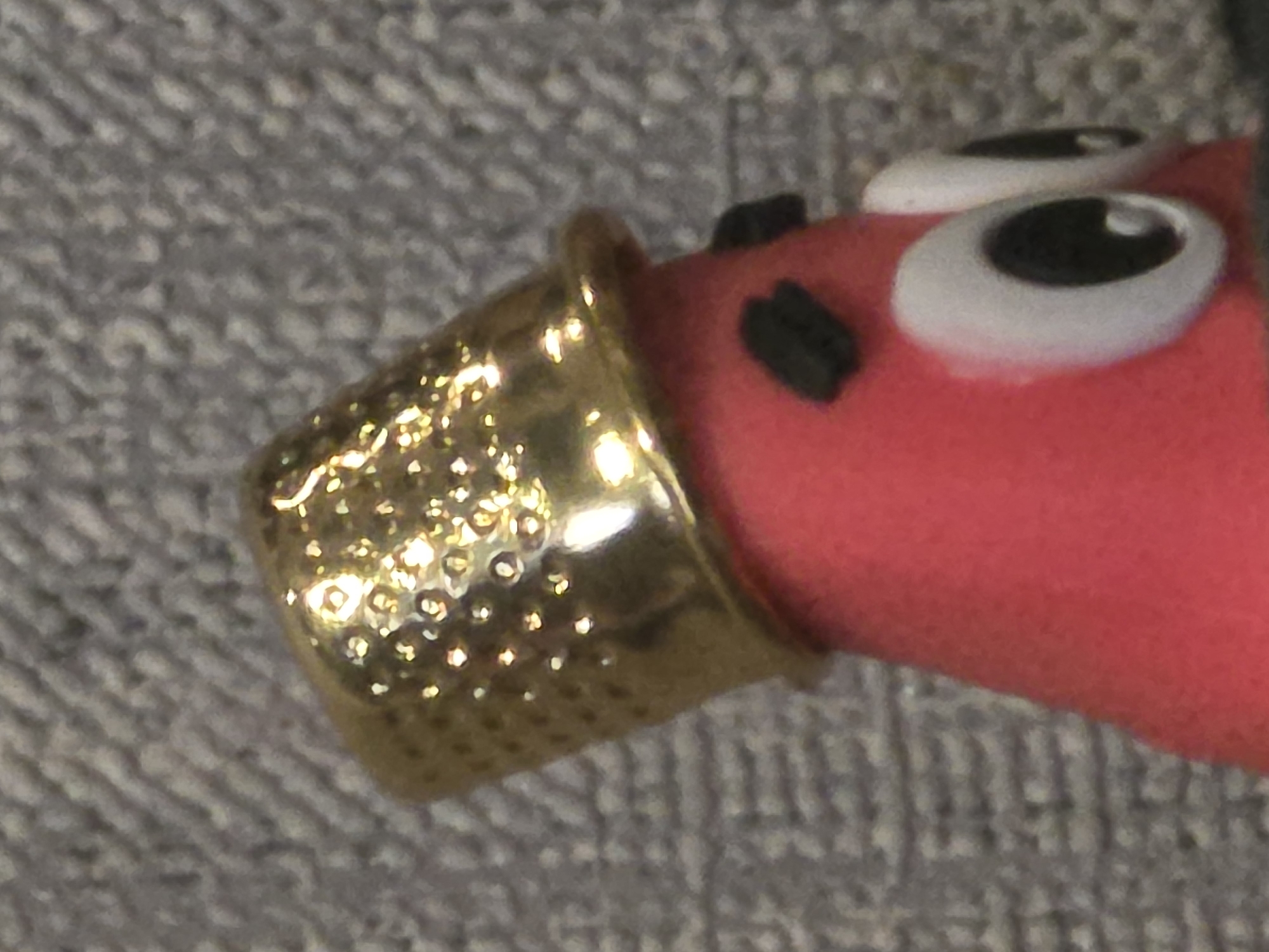 This media file is a close-up, macro photograph of a small, whimsical object. The primary subject is a thimble, made of shiny, golden metal with a textured surface of small indentations. The thimble is positioned at an angle, with its opening facing towards the right and slightly upwards.
Attached to the thimble, or on a surface it's resting on, is a pink, somewhat amorphous object that appears to be a toy or craft creation. This pink object has two large, cartoonish eyes with white sclera and black pupils, giving it a surprised or wide-eyed expression. Above and to the left of the eyes, there are two small, black, rectangular shapes that might represent nostrils or decorative elements. The pink material appears soft and slightly fuzzy.
The background is a textured surface, likely fabric, with a gray and white patterned weave. The lighting in the image is bright, causing significant reflections and highlights on the metallic thimble, which dominate the visual field on the left side of the frame. The focus is sharpest on the thimble and the eyes of the pink object, with the background appearing slightly out of focus. There is no visible text in the image. The overall impression is one of a handcrafted item, possibly part of a larger collection of toys or decorative pieces.