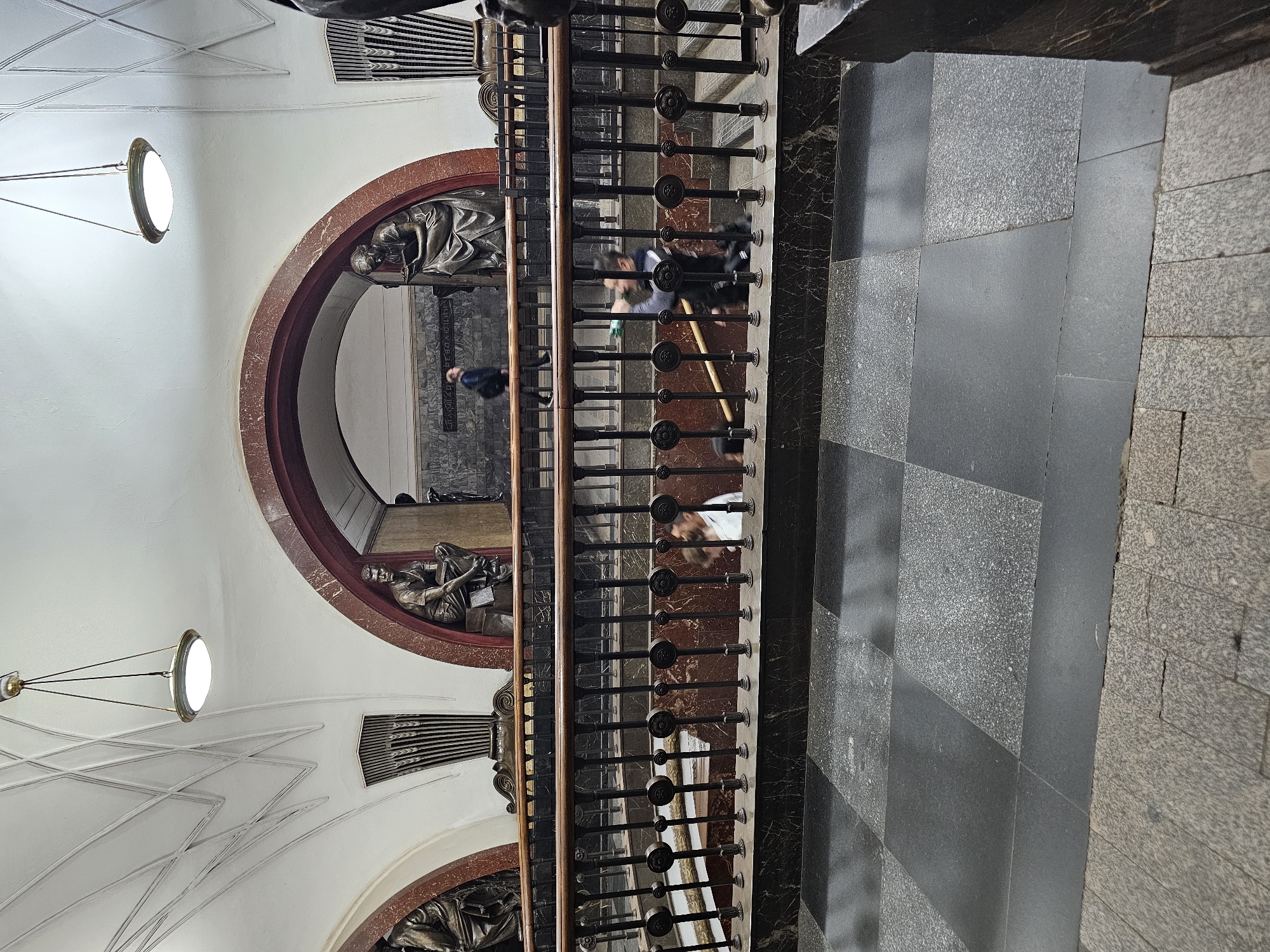 The image shows an ornate staircase inside a Moscow Metro station. The station features classical architectural elements, including arched doorways, large sculptures of people, and decorative railings. Two downward-traveling staircases are visible, separated by a central support structure and a dark metal railing. The floor is paved with large, dark grey tiles interspersed with lighter, speckled grey tiles, creating a checkered pattern. On the upper level, two statues are prominently displayed within arched niches on either side of a central archway. The archway leads to a background area with a marble-clad wall featuring a bas-relief or inscription. Several individuals are descending the stairs. One person in the middle ground is wearing a white shirt and appears to be looking down. Another person in the background is walking away from the camera. In the upper left and right portions of the image, pendant light fixtures with circular shades hang from the white, vaulted ceiling. The overall impression is one of grandeur and historical significance, typical of many older Moscow Metro stations.