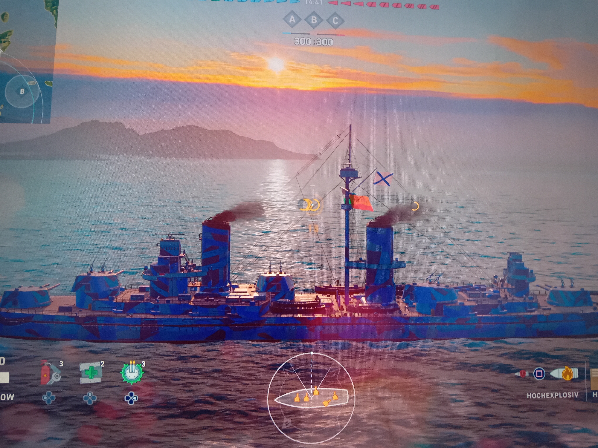 A large warship, likely a battleship or heavy cruiser, dominates the foreground of a video game screen. The ship is painted in a blue and dark blue camouflage pattern, with two main funnels emitting black smoke and multiple gun turrets. It sails on calm, reflective blue water.
The scene depicts a dramatic sunset, with the sun low on the horizon, casting vibrant orange, pink, and purple hues across the sky and reflecting on the water. Silhouetted landmasses, possibly mountains, are visible in the distance under the colorful sky.
Various game interface elements overlay the scene:
- A mini-map in the top-left corner shows a circular area marked "B".
- At the top-center, a timer reads "14:4", with objectives "A", "B", "C", and a score of "300:300".
- On the ship's mast, flags are visible, including a blue and white X-shaped flag and a red/orange flag, alongside two yellow crescent symbols.
- At the bottom-left, icons for ship abilities or consumables show a fire extinguisher (3 uses remaining), a medical kit (2 uses remaining), and a gear-like icon (3 uses remaining). Partial text "OW" is also visible.
- A circular aiming reticle with a ship silhouette and four yellow markers is positioned at the bottom-center.
- At the bottom-right, an ammunition type indicator shows icons for a projectile and fire, with the German text "HOCHEXPLOSIV" (High Explosive) and a partial "H".
The image appears to be a photograph of a screen, showing pixelation and potential reflections.