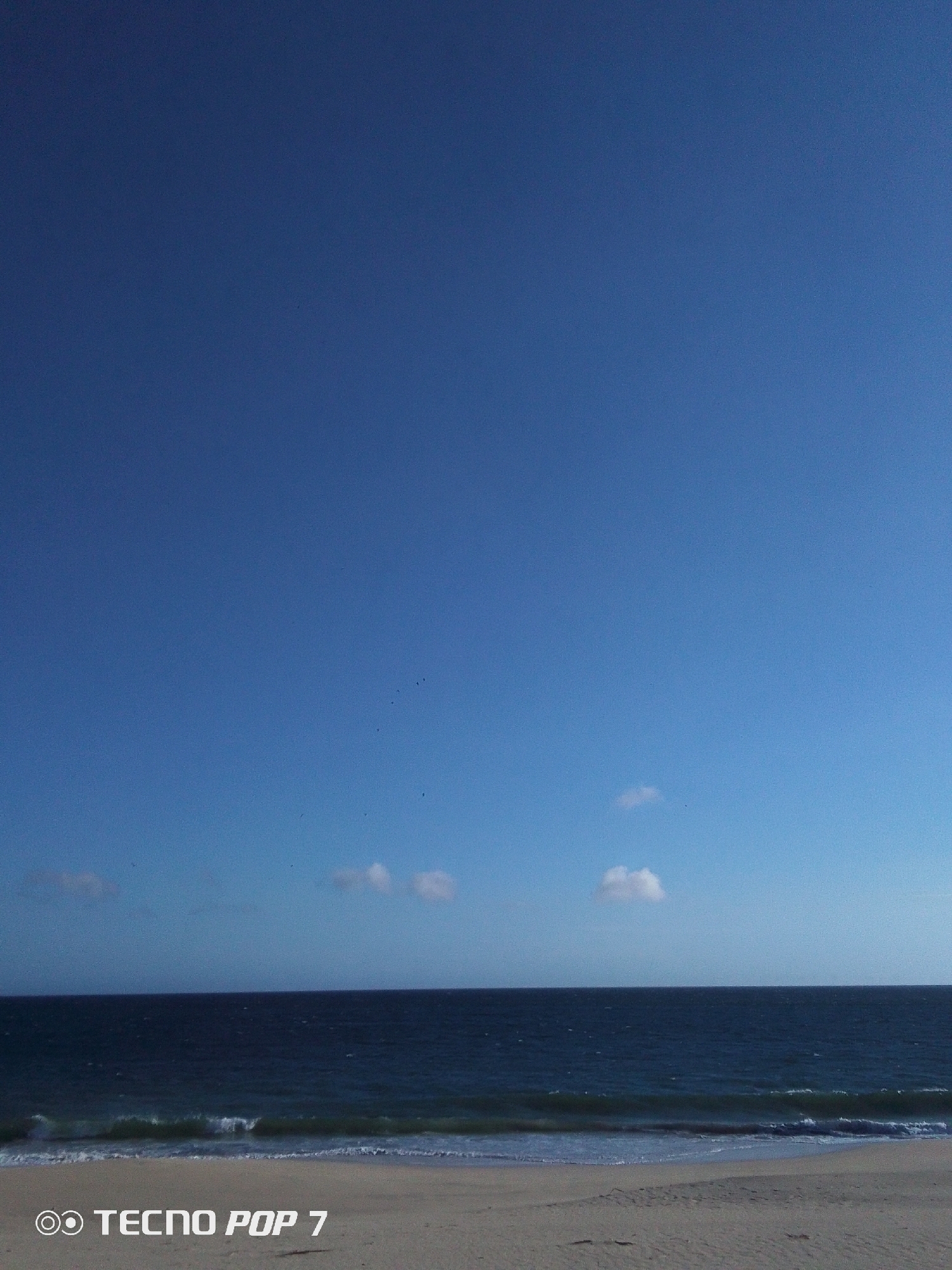 The image depicts a coastal scene at Pedro González, Venezuela. A sandy beach stretches into the foreground, meeting the ocean. Gentle waves are breaking on the shore. The ocean is dark blue and extends to the horizon. The sky is a clear, bright blue with a few small, scattered clouds. The picture was taken with a TECNO POP 7 device.