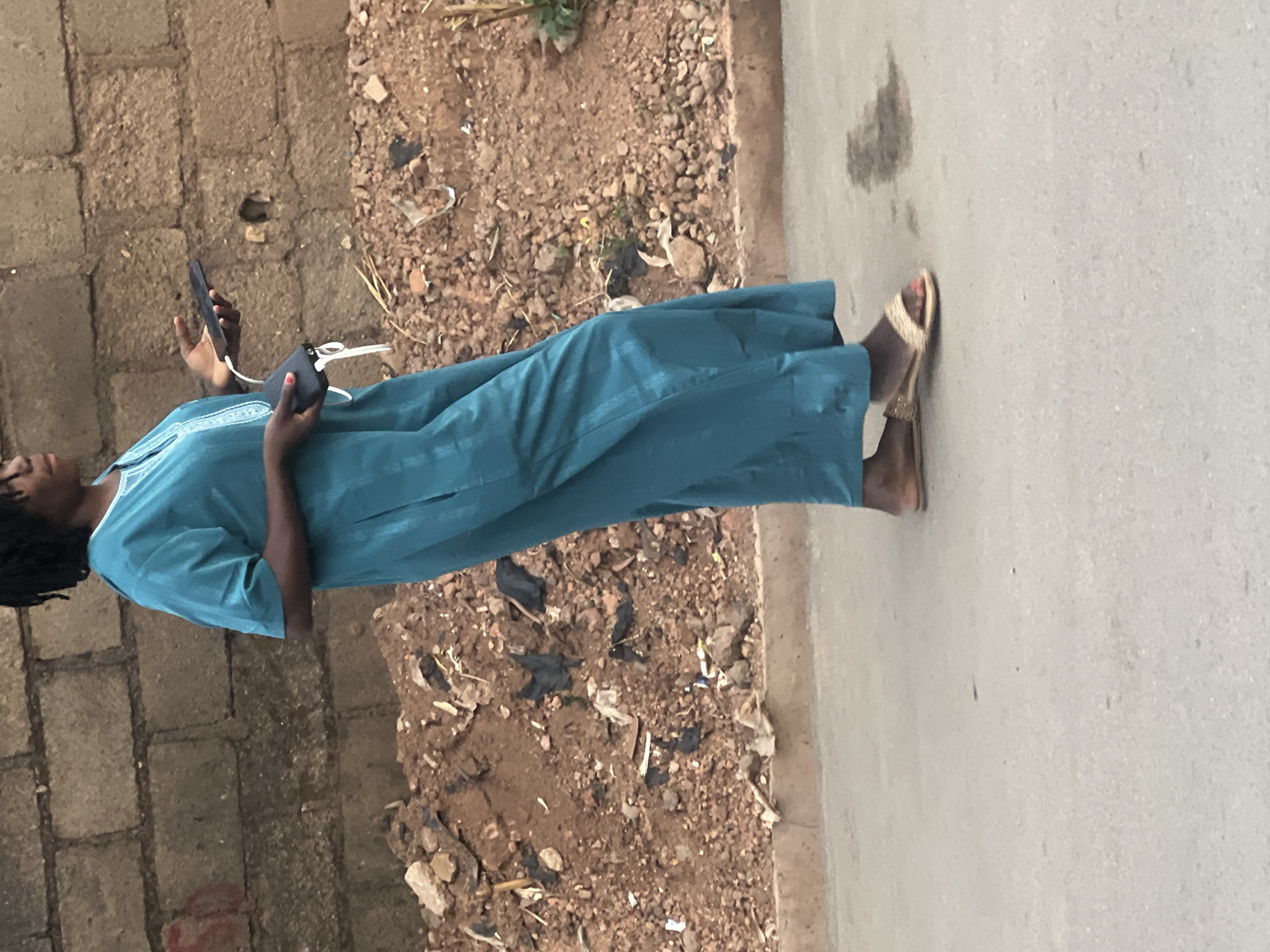 An upright, full-body shot captures a person standing outdoors, engaged with two mobile phones and a power bank. The subject, likely female, has dark skin and dark, possibly braided or dreadlocked hair. She wears a long, flowing, teal-colored kaftan or traditional robe with subtle white detailing around the neckline. On her feet are gold-colored sandals, revealing toes with red nail polish.
In her hands, she holds two dark-colored smartphones. A white cable connects one of the phones to a black power bank, suggesting the phone is being charged. Her gaze is directed downwards at the devices.
The setting appears to be a street or pathway. The person stands on a light grey, paved surface, possibly asphalt or concrete. To her left, there's an unpaved area of reddish-brown dirt, mixed with small stones and pebbles, and scattered bits of litter such as plastic and dry vegetation. A low wall constructed from large, rectangular concrete blocks is visible along the edge of the dirt area.
The lighting is soft, indicating daytime, possibly early morning or late afternoon, with no harsh shadows. The overall scene depicts a mundane moment of a person using modern technology in a typical outdoor, semi-urban environment. No specific text is visible in the image.