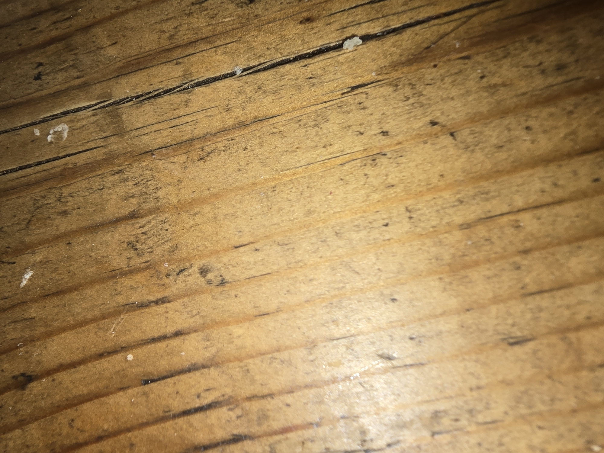 This is a close-up, low-angle, overhead shot of a weathered wooden surface. The wood appears to be planks or boards laid parallel to each other, running diagonally from the bottom left to the top right of the frame. The grain of the wood is visible, with darker lines accentuating the natural pattern. The surface is a warm, medium brown color, with variations in tone. There are numerous black marks and smudges scattered across the wood, suggesting wear and tear. Some small, white specks are also visible, possibly dust, debris, or residue. The lighting is dim, with shadows highlighting the texture of the wood and creating a slightly textured, rustic appearance. The image does not contain any people or identifiable objects beyond the wooden surface itself. There is no information to place this image in Ulan-Ude, Russia.