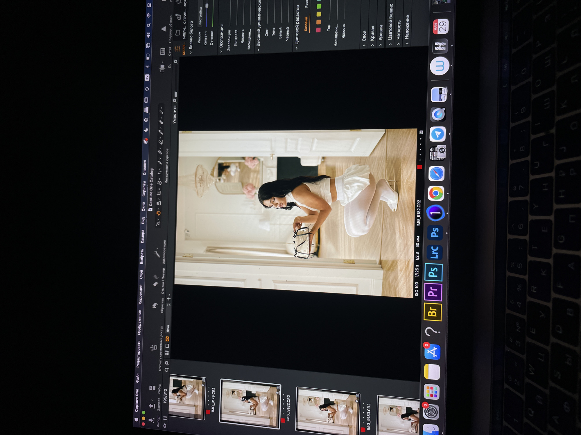 A person with long black hair is posing on the floor for a photo shoot. She is wearing a white outfit and holding glasses in her hand. The photograph being edited is displayed on a MacBook Pro screen, utilizing Capture One software. Various image editing tools and file thumbnails are visible on the screen. The laptop is placed on a desk with a lit keyboard.