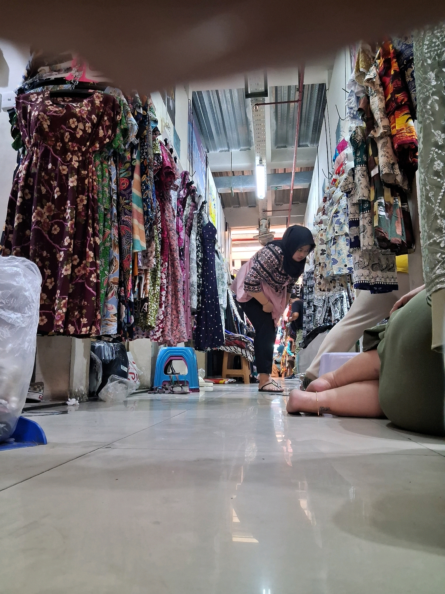 A low-angle, wide shot captures a bustling market stall scene in Surakarta, Indonesia. In the foreground, the tiled floor reflects the overhead lighting and the merchandise displayed. To the left, racks of colorful dresses with floral and geometric patterns hang, with a large plastic bag and a blue plastic stool visible near the base of the racks. A woman in a hijab and a pink and patterned blouse is bent over, inspecting items at a stall on the right. Her dark pants and sandals are visible. Further in the background, other shoppers are present, and additional stalls with clothing are visible, extending down a narrow aisle. The overall impression is one of a lively and crowded marketplace.
