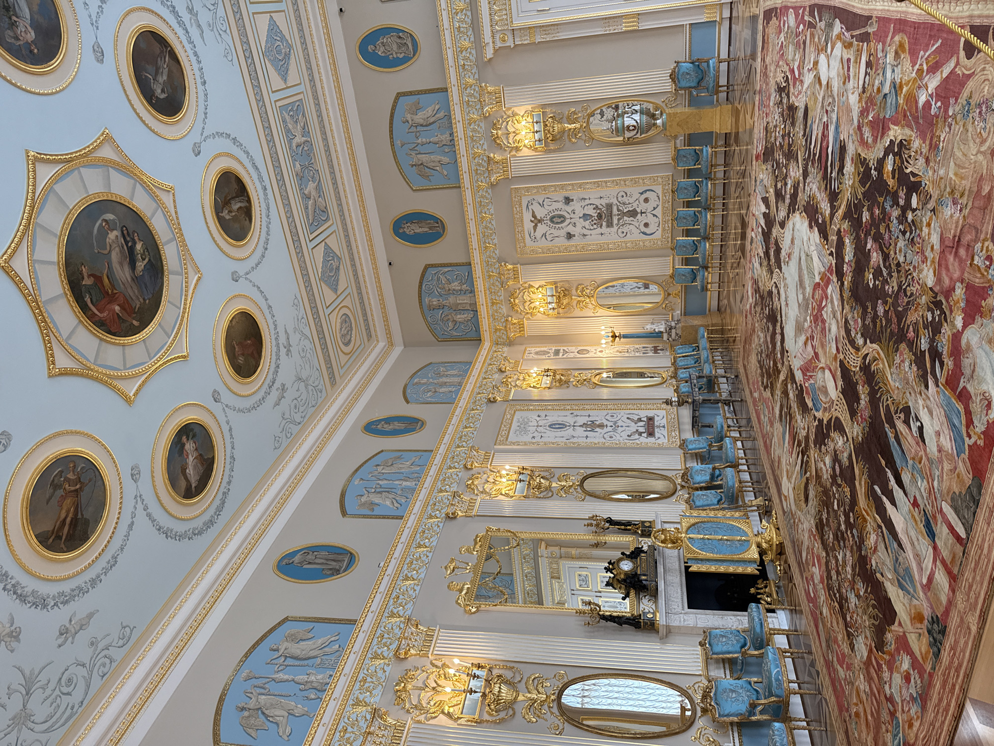 This is an interior view of the drawing room in the Catherine Palace in Pushkin, Russia. The room is elaborately decorated with gilded ornamentation and painted ceilings. A large, intricate carpet covers the majority of the floor. Along the walls are rows of light blue upholstered chairs with gilded frames, and several ornate mirrors and sconces. In the background, a fireplace with a mantelpiece and a clock can be seen. The ceiling is adorned with multiple round medallions featuring painted figures and decorative stucco work.