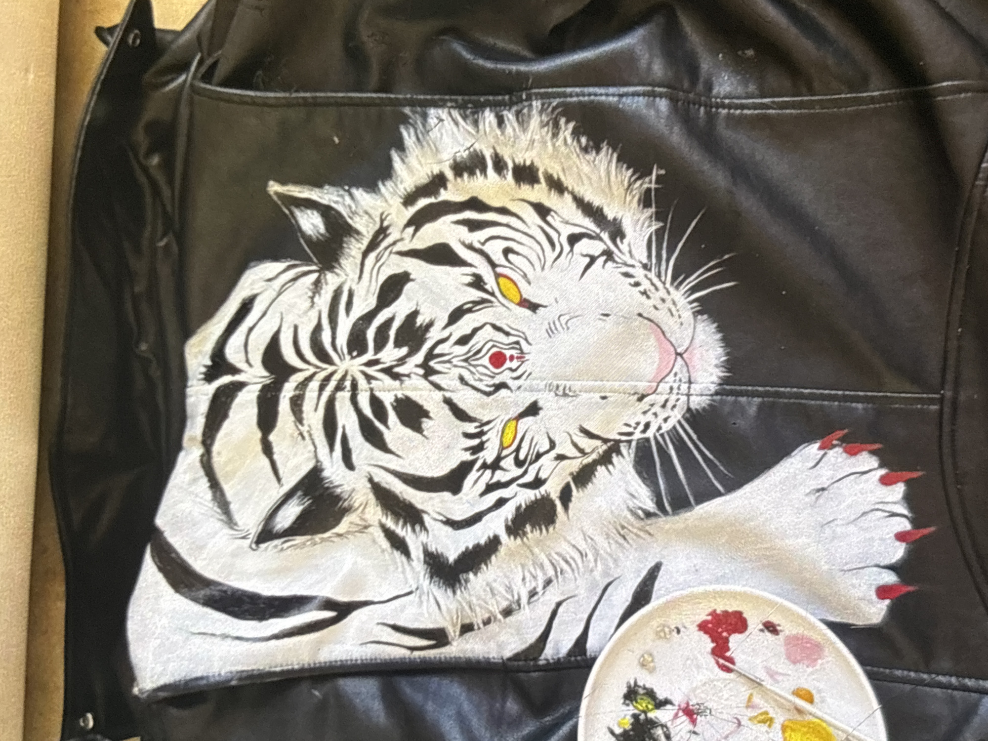 The image shows a close-up of a black leather jacket with a painted white tiger on the back. The tiger is depicted with black stripes, yellow eyes, a pink nose, and red claws. A small white plate with colorful paint splatters is visible in the lower left corner. The jacket is lying flat, and the background is a neutral, textured surface. There is no visible information within the image to determine its location in Orihuela, Spain.