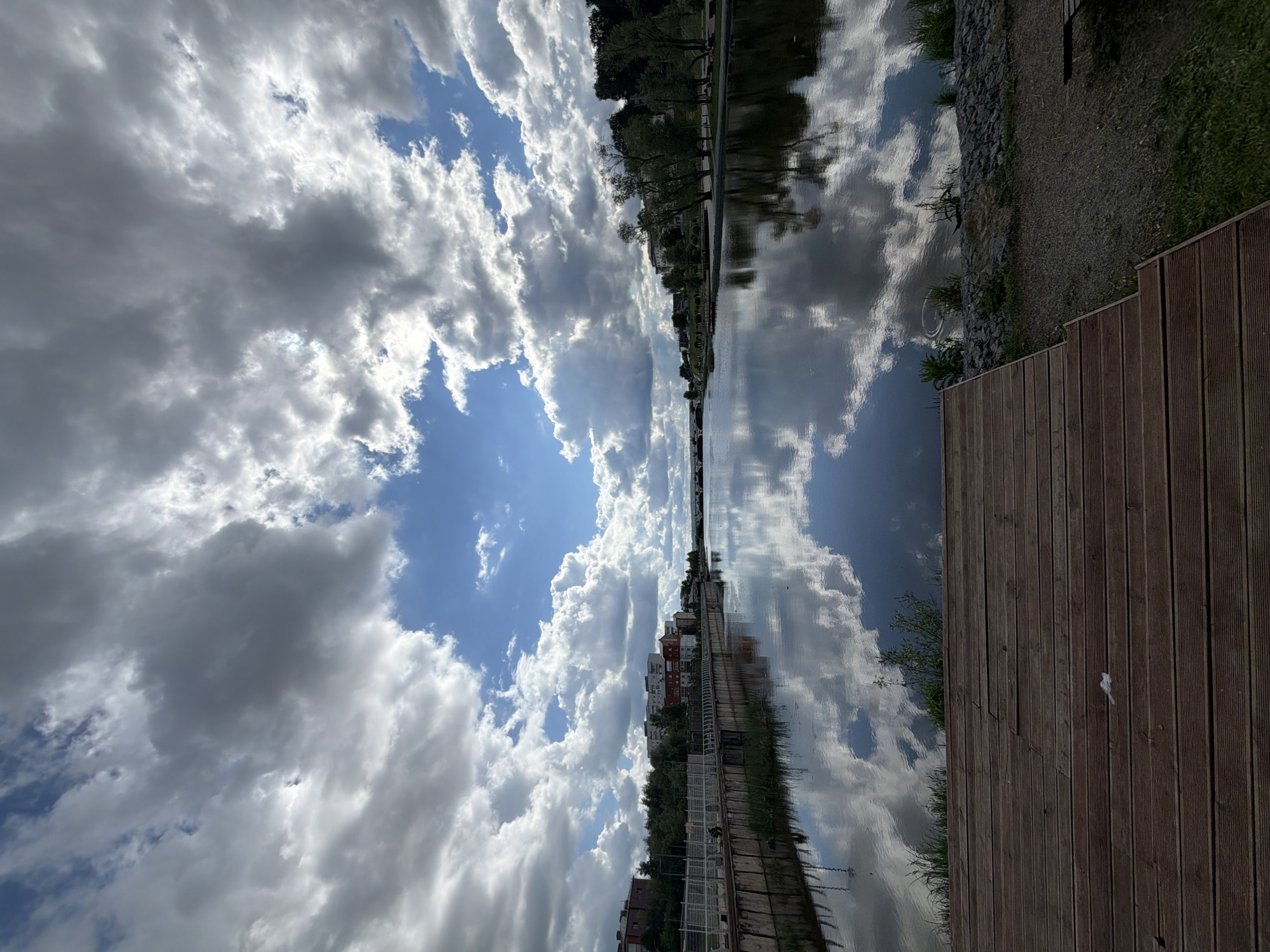 The image depicts a scene along the Votkinsk Reservoir in Votkinsk, Russia, showing a waterfront promenade. The sky is partially cloudy with a mix of cumulus and stratocumulus clouds, some reflecting in the still water of the reservoir. Buildings are seen along the opposite shore, some appearing residential. Trees line the shoreline. A wooden deck extends along the edge of the water. The image is taken from an off-camber angle.