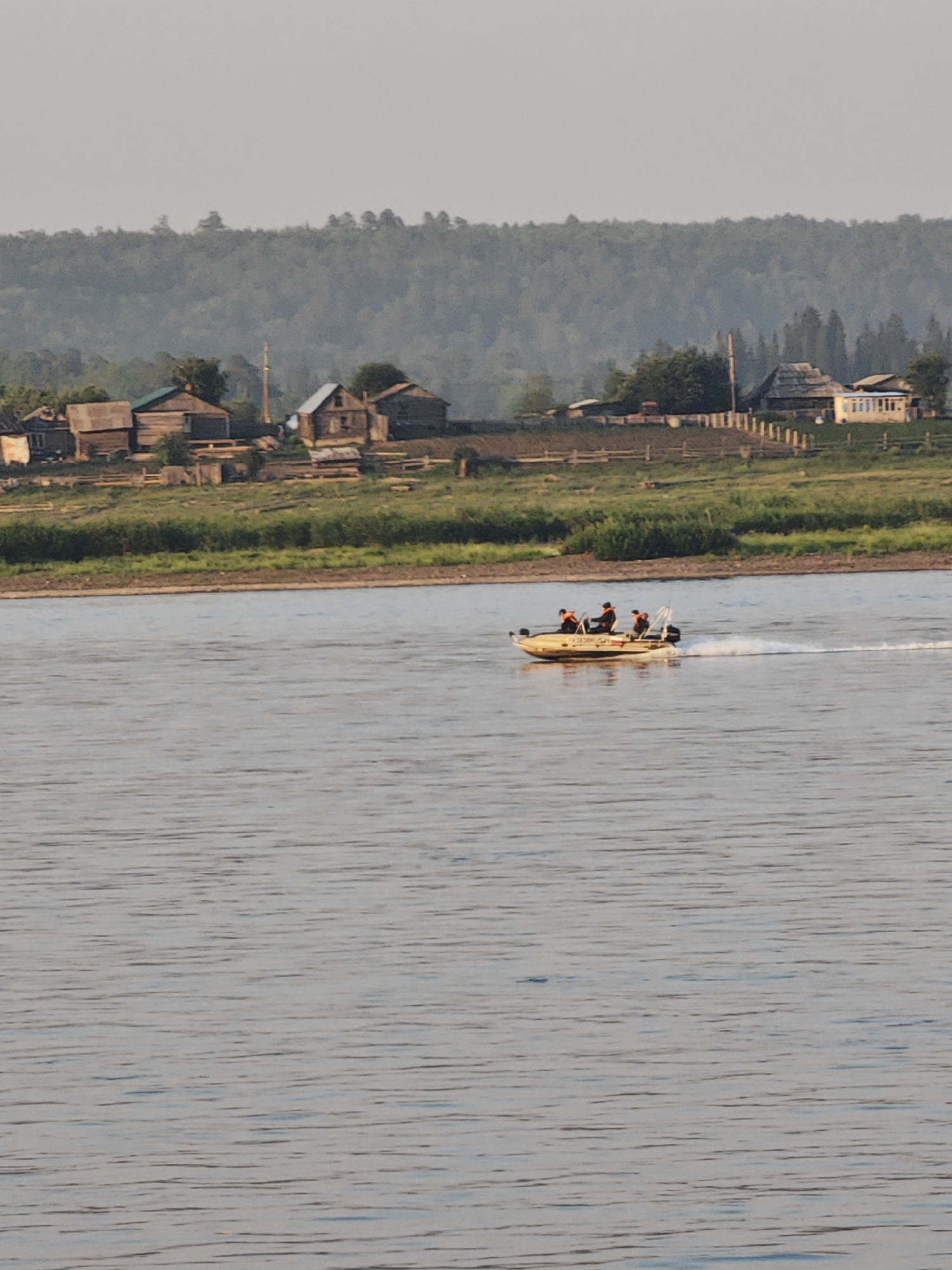 A motorboat with three occupants is traversing a wide, calm river. The boat is light-colored and appears to be moving at a moderate speed, leaving a wake behind it. The occupants are wearing orange life vests. In the background, a distant shoreline features a cluster of rustic wooden houses and buildings, suggesting a rural or semi-rural settlement. Beyond the settlement, a dense forest covers a rolling hillside. The sky above is overcast with a soft, hazy light. The overall scene depicts a tranquil day on a river in a natural setting with evidence of human habitation.