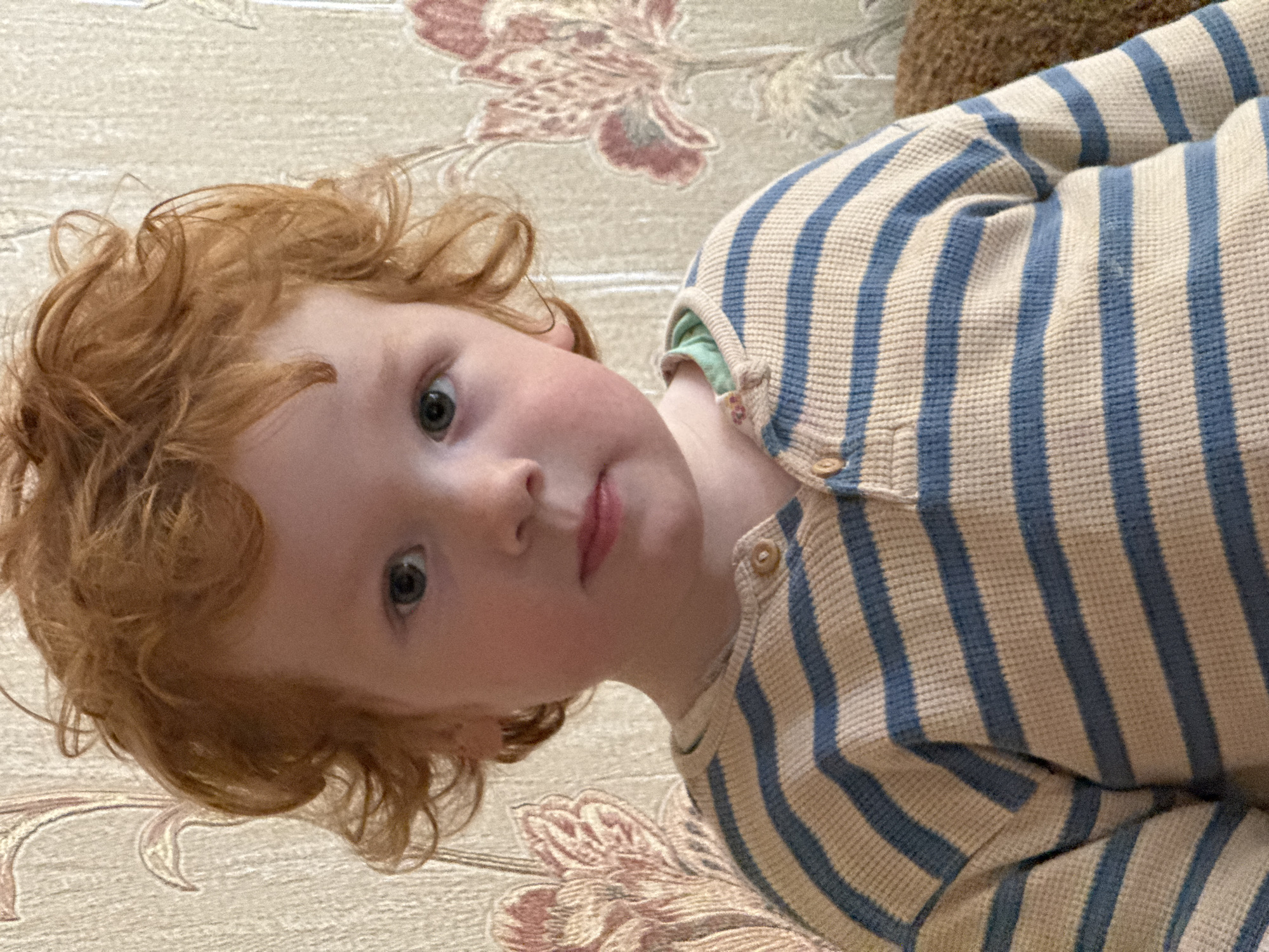 A close-up portrait shows a young child with fair skin, reddish-blonde curly hair, and light-colored eyes. The child is wearing a striped shirt of blue and beige, with button closures. The child's gaze is directed slightly upwards, and their expression is somewhat neutral. The background reveals a patterned surface, possibly a rug or upholstery, with floral designs in beige and reddish-brown hues. The overall setting and style of the image do not provide evidence to suggest a location in Saint Petersburg, Russia, and based solely on the image, it is impossible to determine the city where the photo was taken.