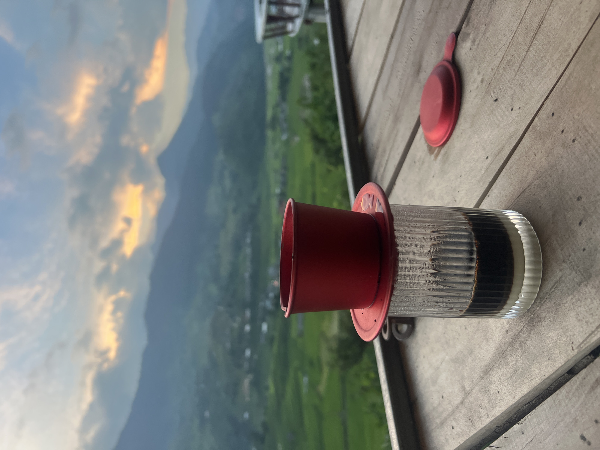 A close-up shot features a Vietnamese coffee preparation with a red metal filter atop a clear glass filled with condensed milk and coffee. The glass sits on a wooden table, with a red lid resting nearby. In the background, a lush green valley with terraced rice fields stretches towards misty mountains under a cloudy sky during sunset. The scene is set in Tu Le, Vietnam, a region known for its picturesque rice terraces.