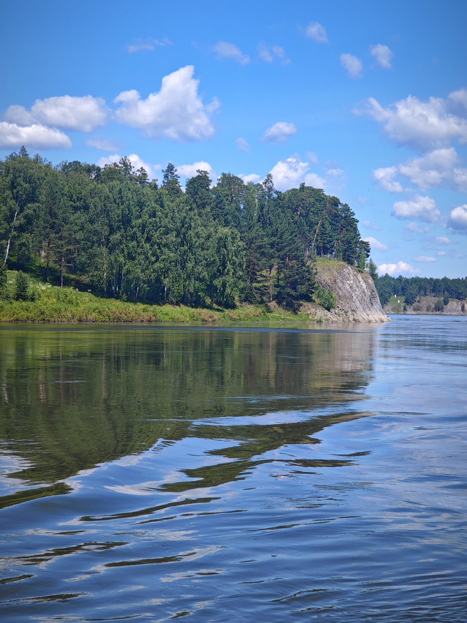 The image depicts a serene natural landscape under a bright blue sky dotted with fluffy white clouds. A wide, calm river dominates the foreground, its surface reflecting the lush green trees and rocky cliffs that line its bank. The vegetation is dense, featuring a mix of deciduous and coniferous trees, with a steep, rocky incline on the right side of the riverbank. The water exhibits gentle ripples, suggesting a slight current or the passage of a boat. The scene appears to be in a rural or natural setting, possibly a park or a nature reserve, with no visible human presence or man-made structures apart from a distant, small white tower on the right bank. The overall impression is one of tranquility and natural beauty. The specific location is identified as being in the city of Savino, Russia.