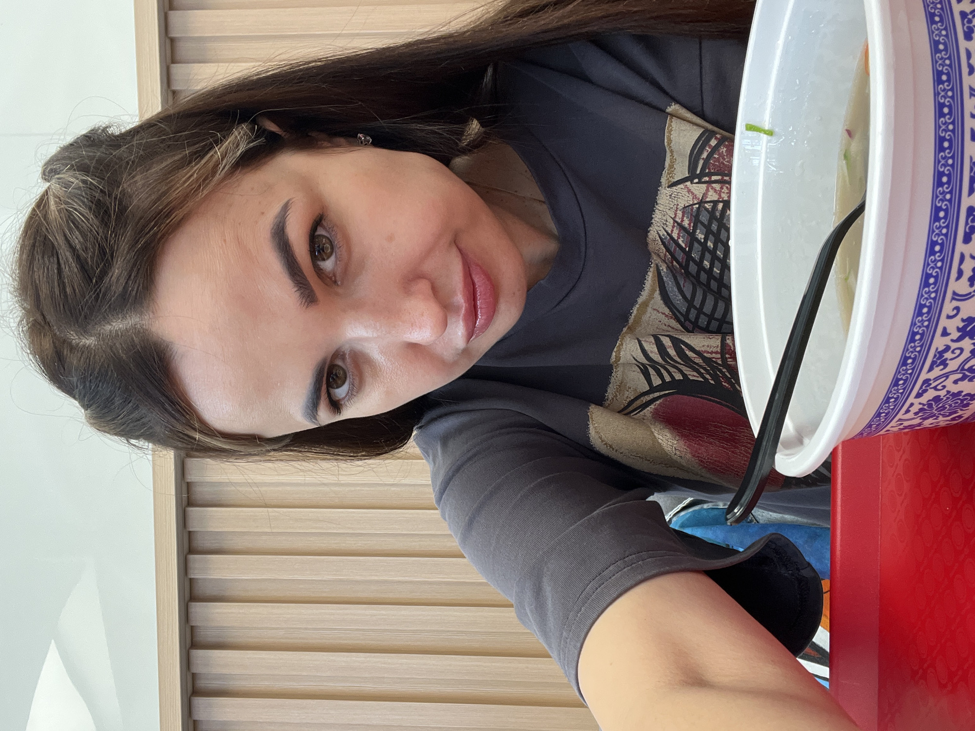 A medium shot captures a young adult woman from the shoulders up, facing the camera with a slight tilt of her head. She has long, dark brown hair styled with two small top knots, and her eyes are brown. She is wearing a dark grey t-shirt with a graphic print featuring abstract shapes in red, tan, and black. A black plastic spoon rests in a white bowl with blue decorative trim, which is filled with a light-colored liquid and visible vegetables, suggesting a meal. The background consists of vertical wooden slats, likely a wall or partition, in a light natural wood tone. The woman is seated at a red table. The overall environment appears to be an indoor dining area.