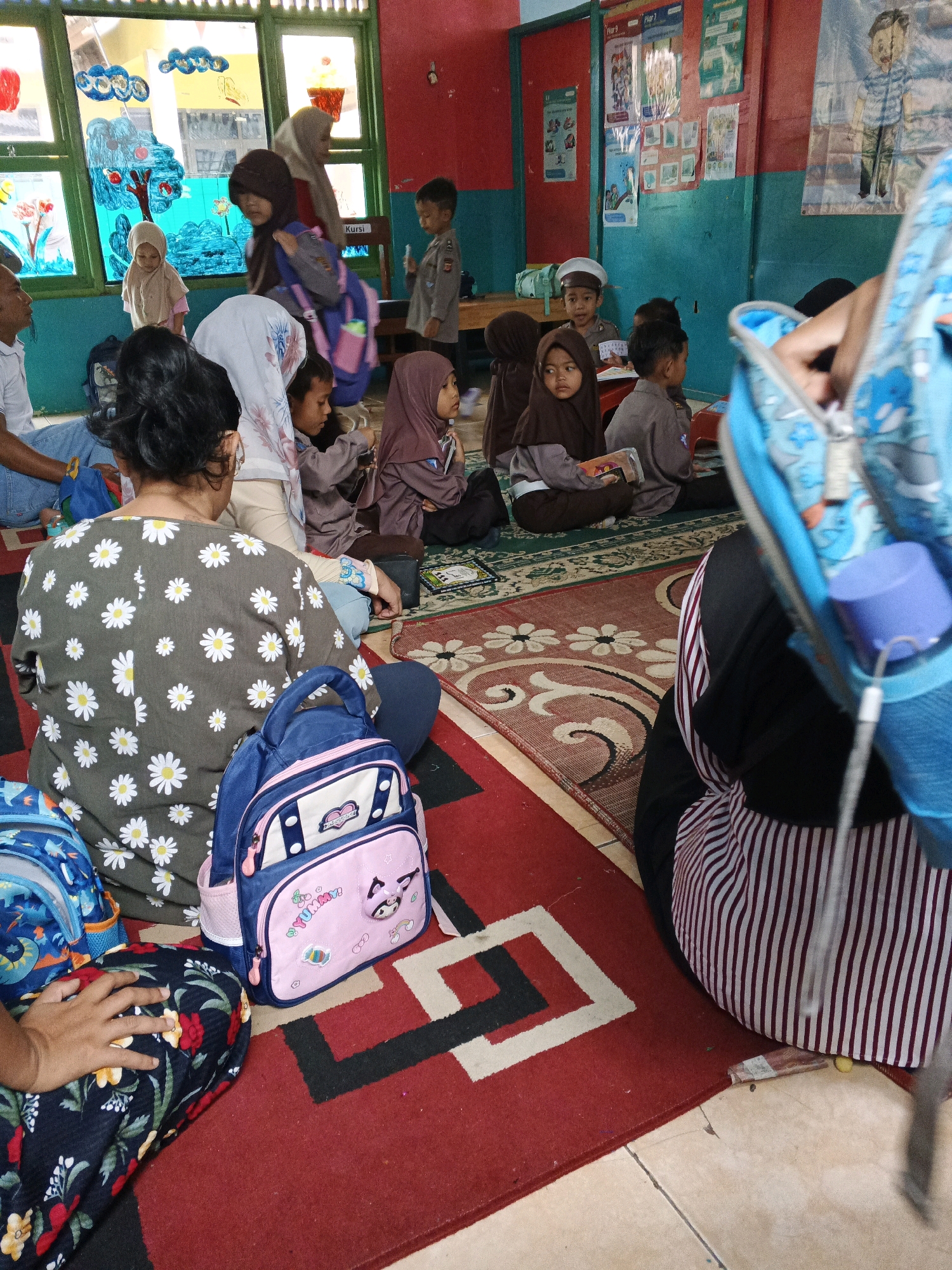 The image depicts a group of children and adults seated on the floor, likely in a classroom setting, in Warungkiara, Indonesia. The children are predominantly wearing uniforms and hijabs, suggesting a school or religious educational environment. They are engaged in various activities, with some appearing to listen, while others hold books or belongings. Backpacks are visible throughout the scene, with one child in the foreground holding a pink and blue backpack. The room features colorful wall decorations and artwork, possibly created by the children. Sunlight streams through a window, illuminating the space. The overall atmosphere suggests a lesson or gathering in progress.