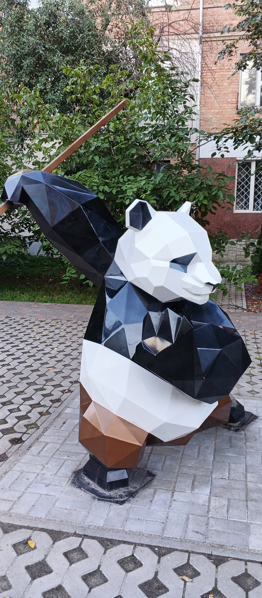 The image shows a low-poly sculpture of a panda in a public space, likely a park or a courtyard, in the city of Smila, Ukraine. The sculpture is made of geometric shapes and is primarily white, black, and brown. It is positioned on a paved surface composed of interlocking concrete blocks. Behind the sculpture, there are trees with green foliage and a brick building with windows. A wooden beam extends diagonally from the upper left of the frame, partially obscuring the trees and the sculpture's arm. The ground is tiled with two different patterns of concrete pavers, with some fallen leaves visible. The overall lighting suggests an outdoor daytime scene, possibly overcast given the diffused light. There are no people visible in the image.